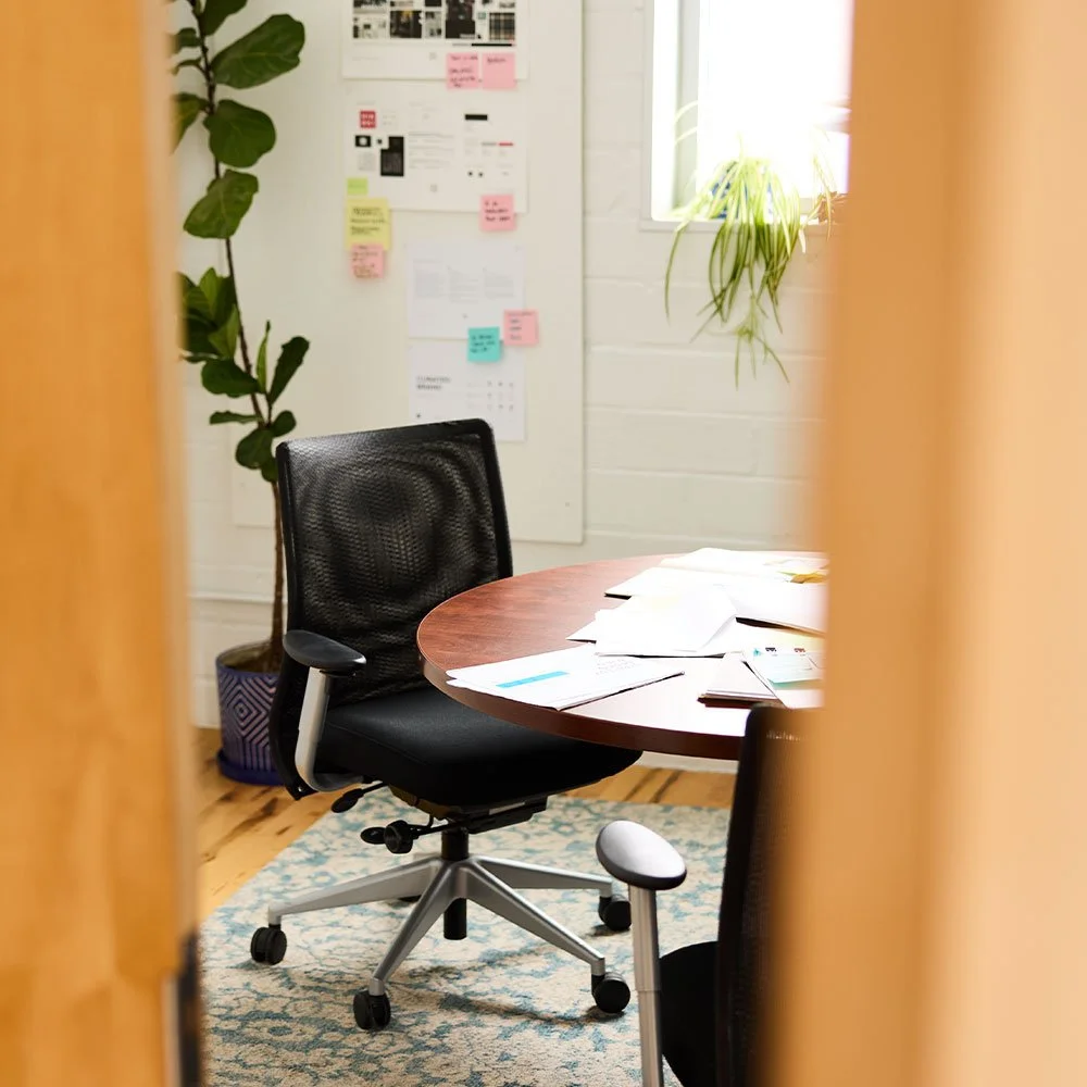 A view through a doorway of a round wooden meeting table with design papers and a black mesh office chair.