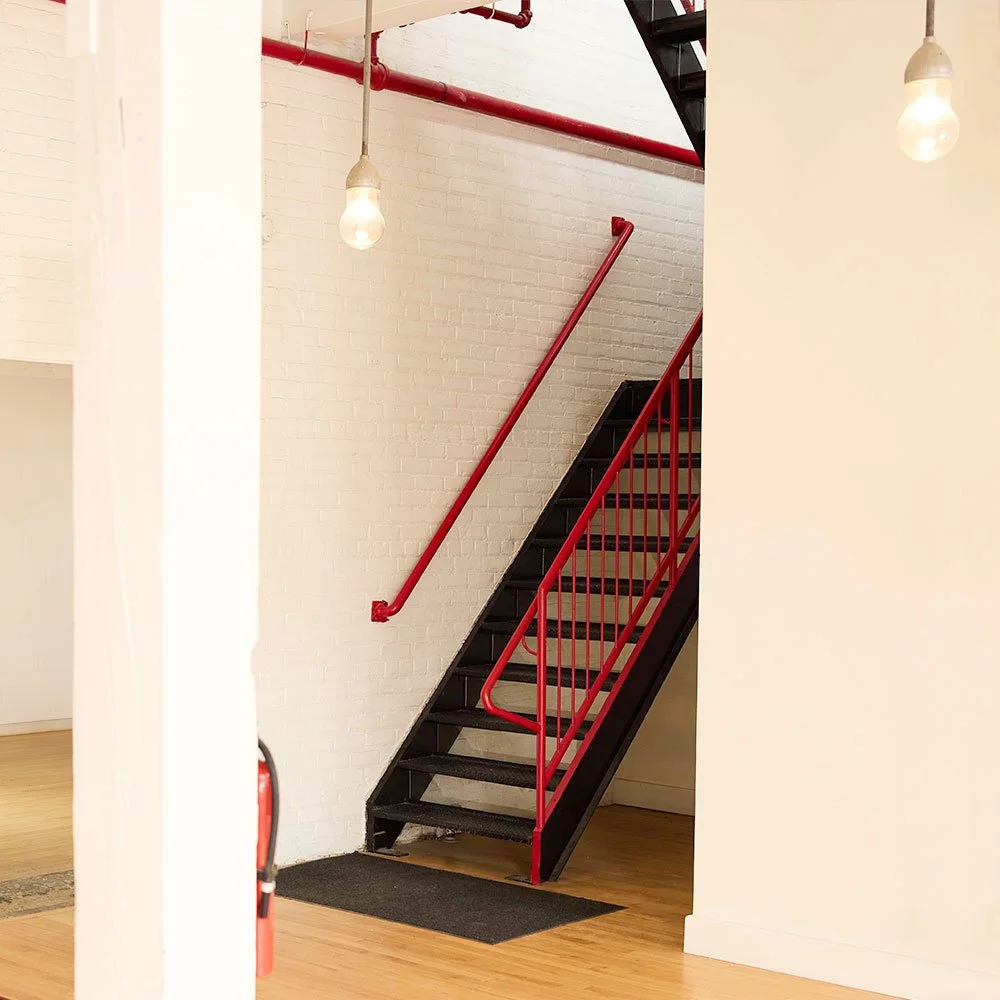 A black industrial staircase with vibrant red railings sits against a white brick wall in a brightly lit studio.