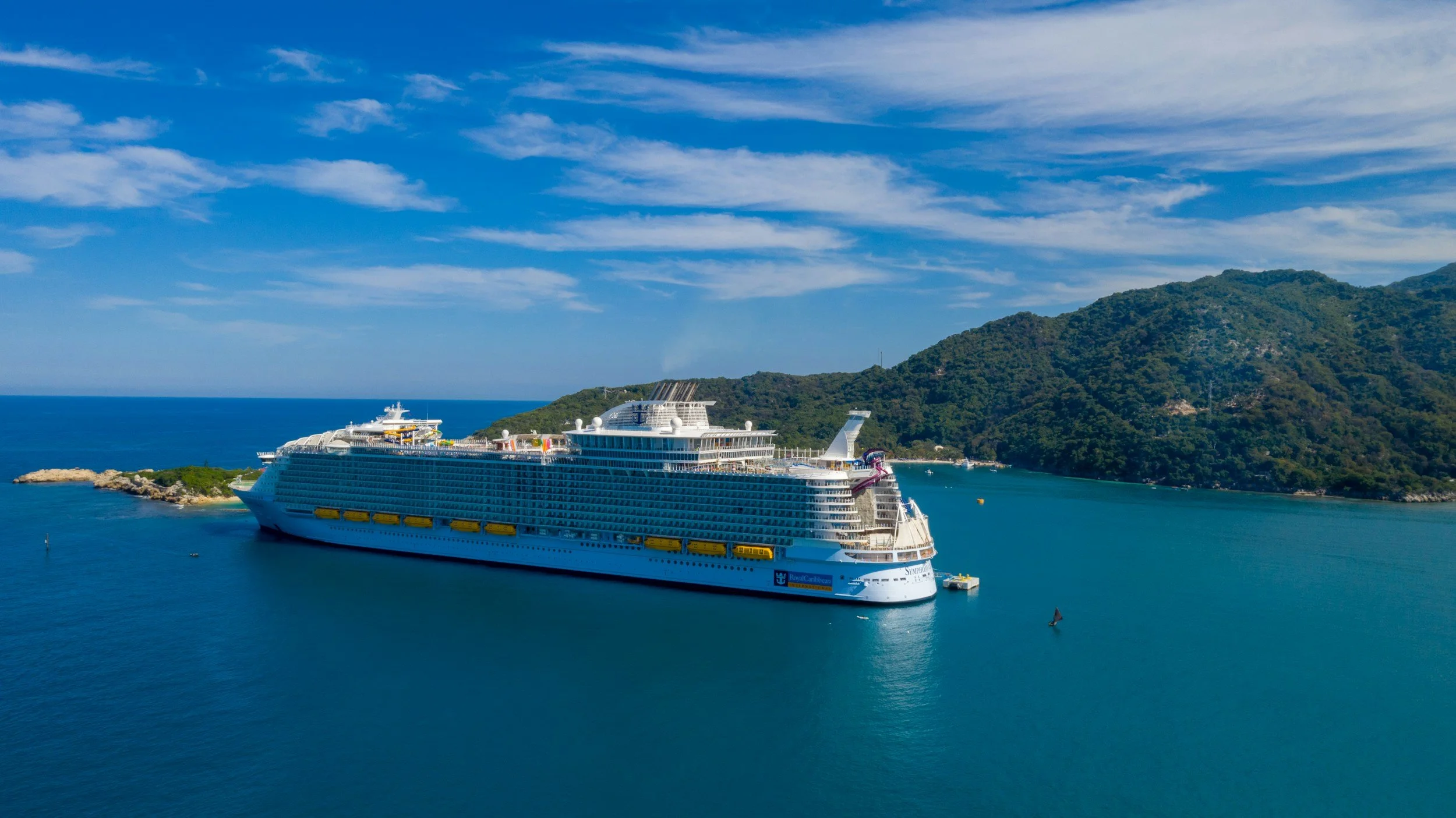A large cruise ship sailing in a calm blue ocean with green mountainous land in the background, under a partly cloudy sky.