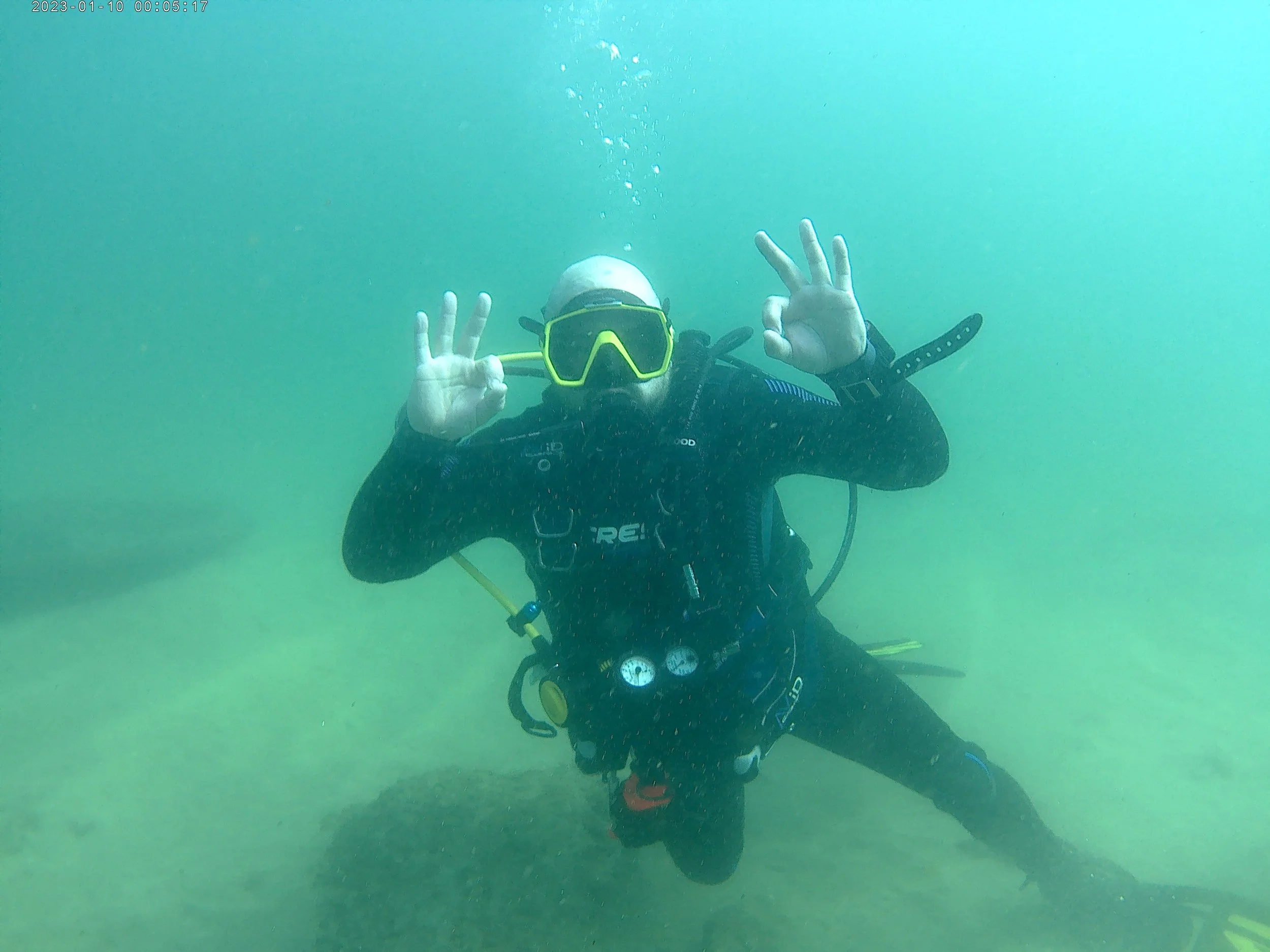 An underwater diver wearing a mask and wetsuit making an OK hand sign and peace sign, with the ocean floor visible in the background.