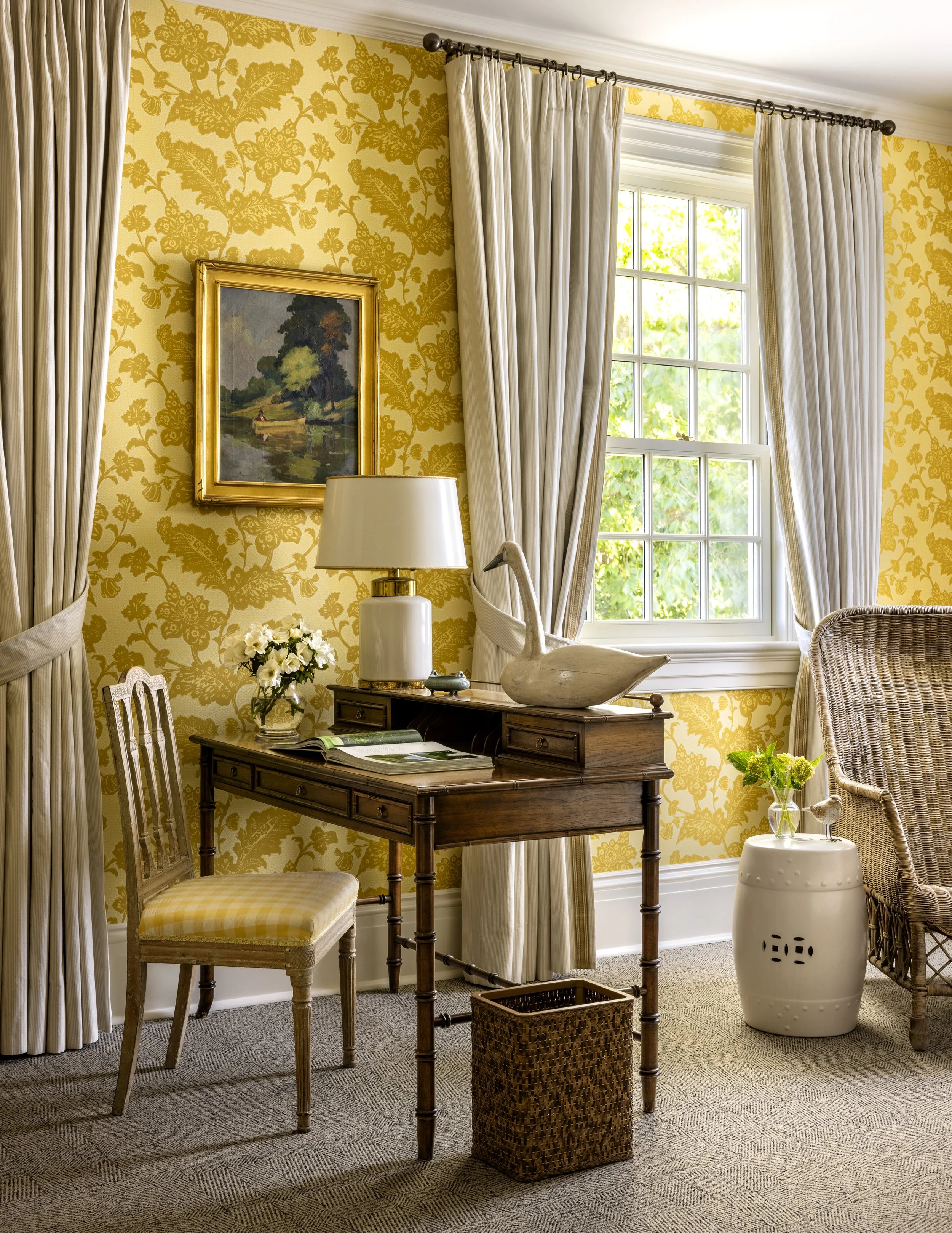 A cozy living room corner with cream-colored floral wallpaper, cream curtains on a window, a landscape painting in a gold frame, a white table lamp, a wooden table with a decorative swan sculpture, and a wicker chair with a yellow checked cushion.