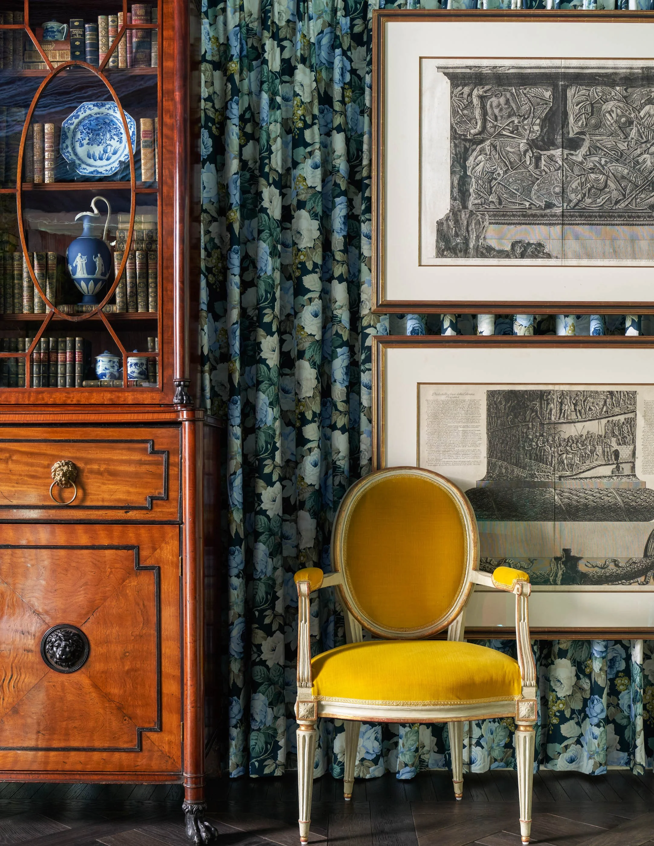 A room with a wooden bookshelf filled with books and decorative blue and white porcelain vases. There are floral curtains in shades of blue, white, and green. A framed black-and-white drawing or etching hangs on the wall. In front of the artwork, the