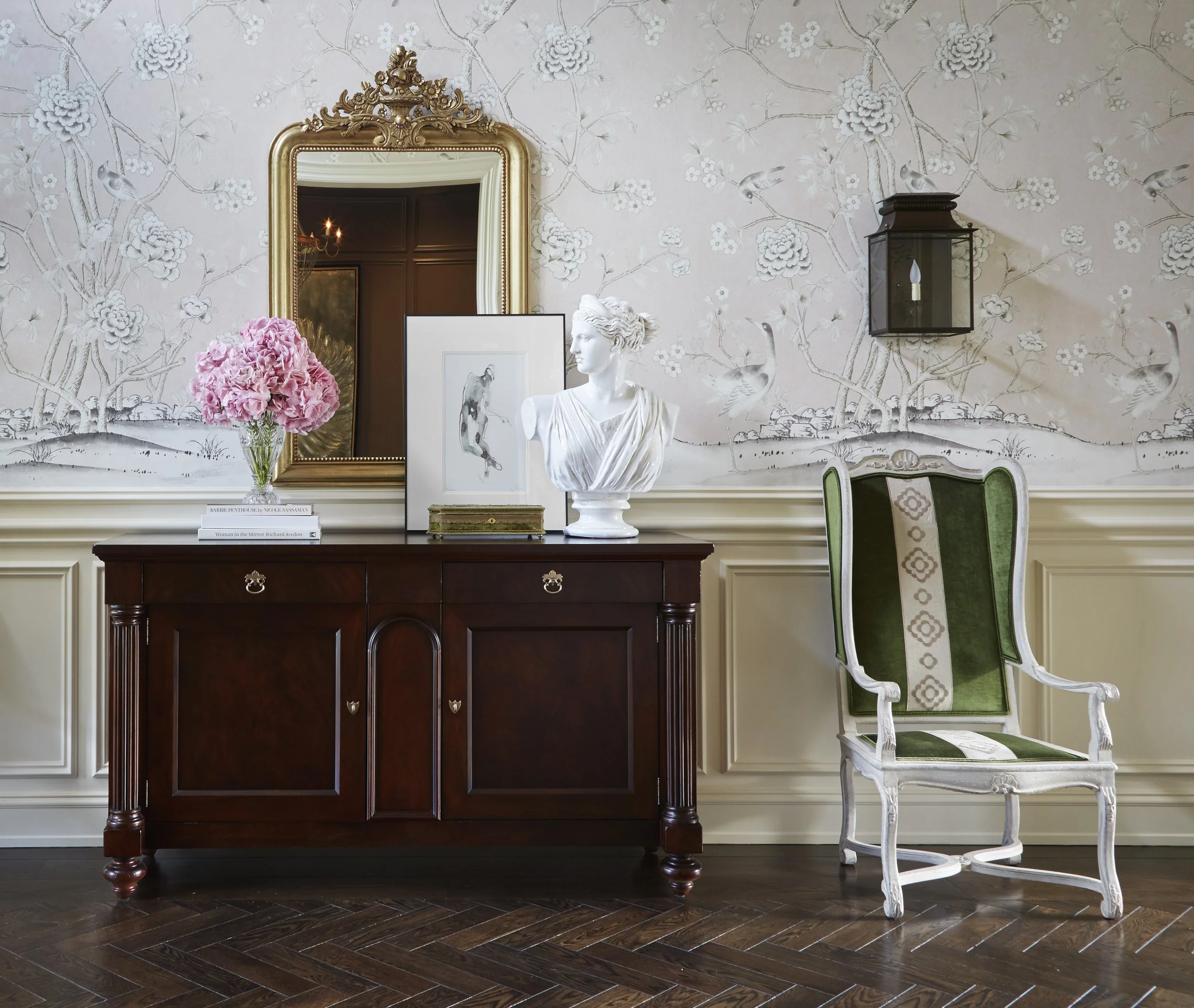 Elegant living room corner with a dark wooden console table, a gold-framed mirror, a classical white bust sculpture, a pink hydrangea flower arrangement, framed artwork, vintage green and white chair, floral wallpaper with birds, and dark hardwood fl