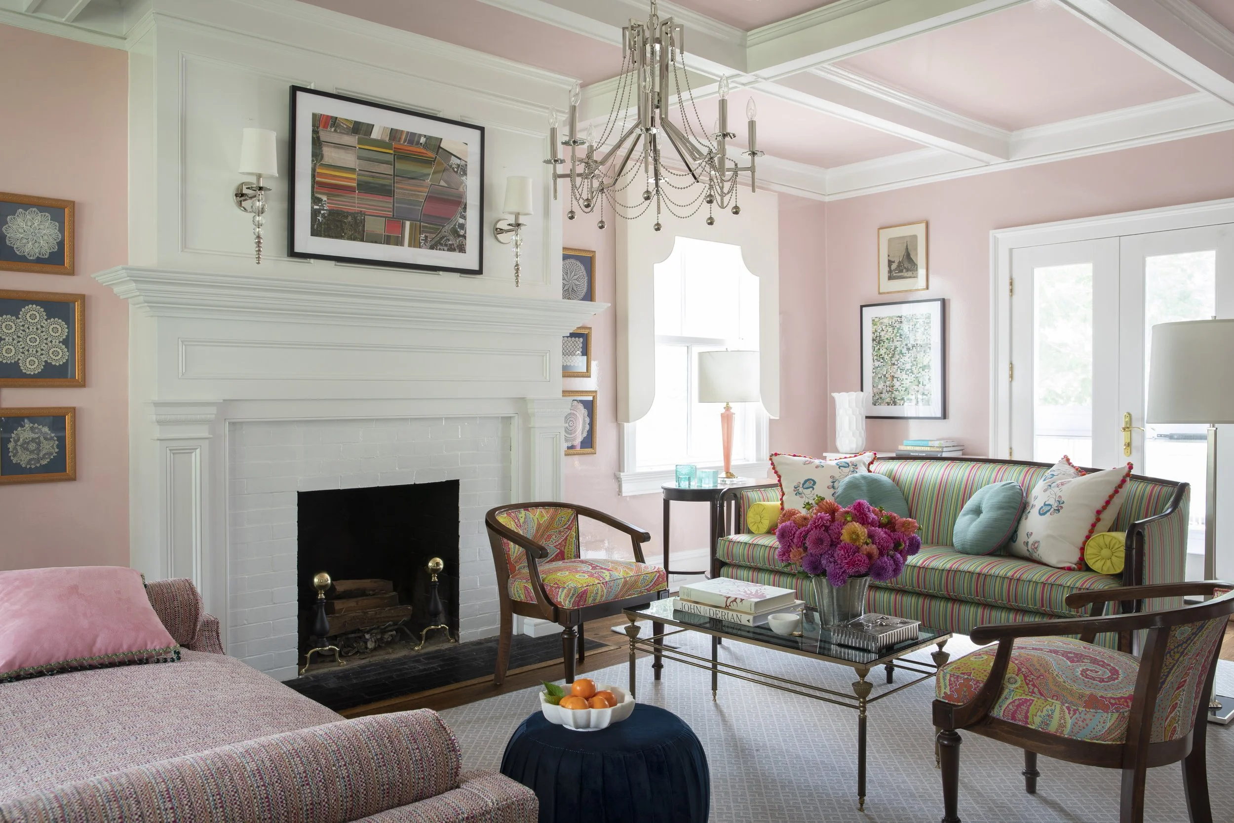 A cozy living room with pink walls and white trim, featuring a white fireplace with a tall mantel, surrounded by framed artwork. There is a colorful striped sofa with decorative pillows, a glass coffee table with a stack of books and a flower arrange