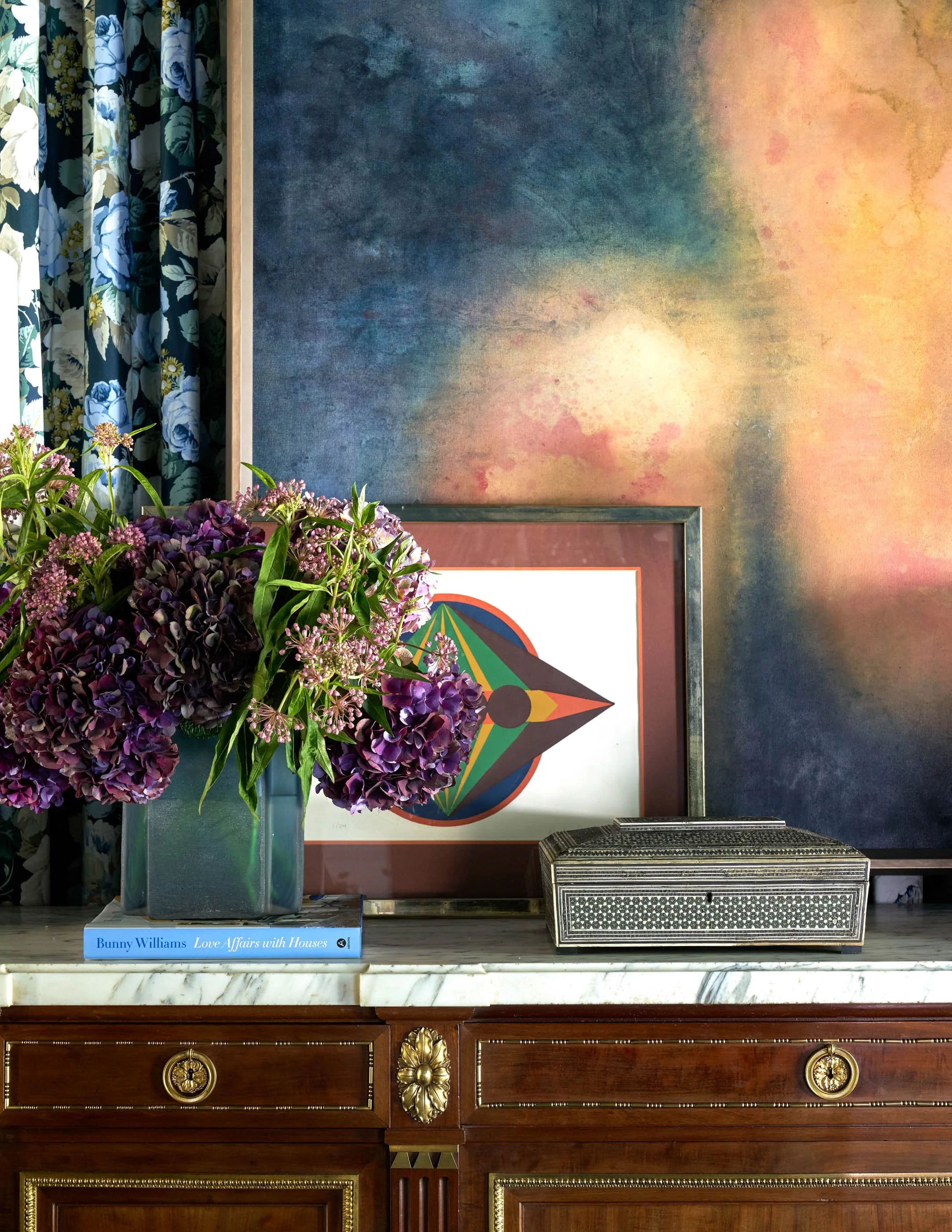 A blue vase with purple hydrangea and other mixed flowers sitting on a marble surface. Behind the flowers, there is a framed colorful geometric art piece on a wall with a mottled blue, yellow, and pink background. To the right of the flowers, there's