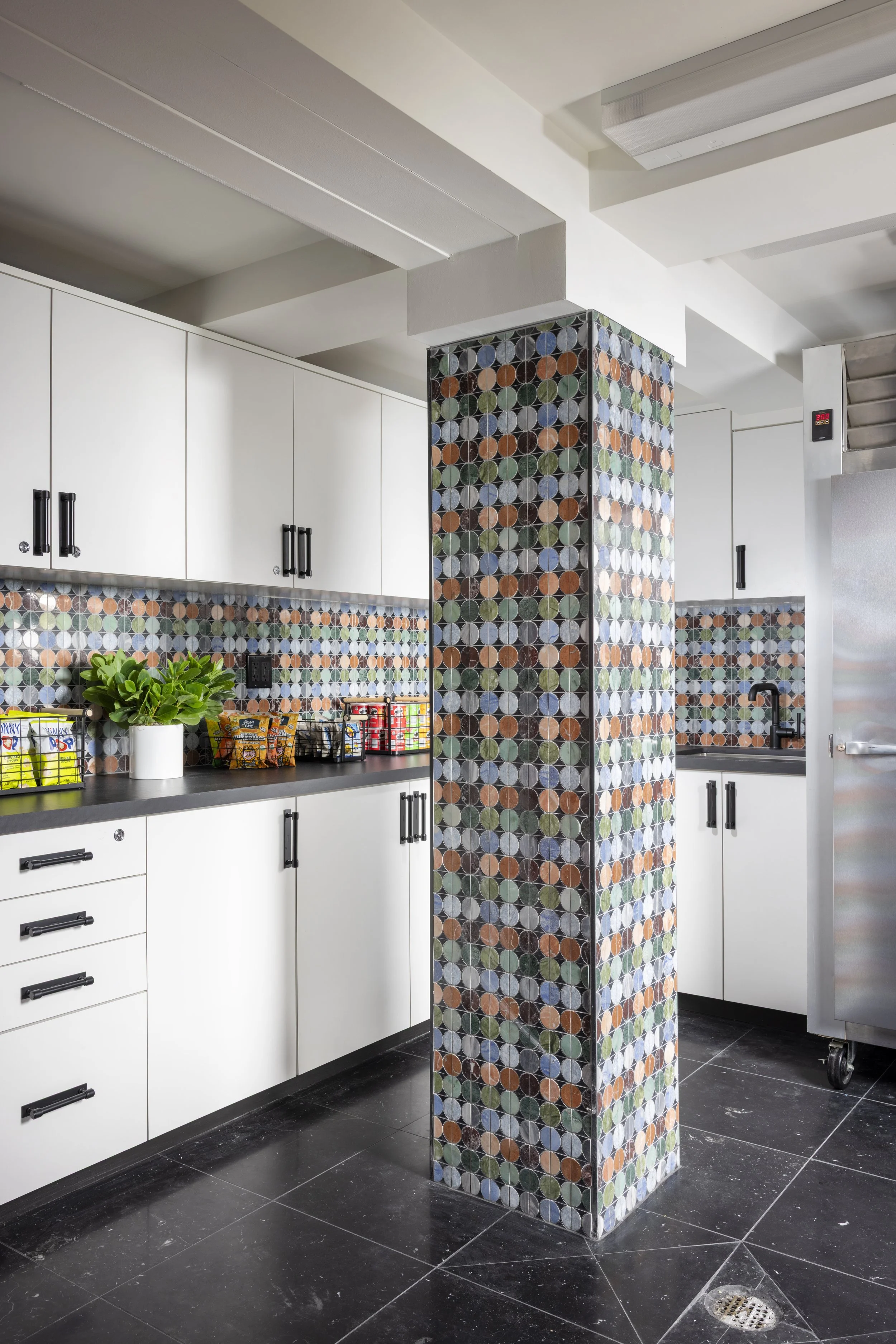 Modern kitchen interior with white cabinets, black handles, a colorful tile pillar, black floor tiles, a plant, and some snack bags on the counter.