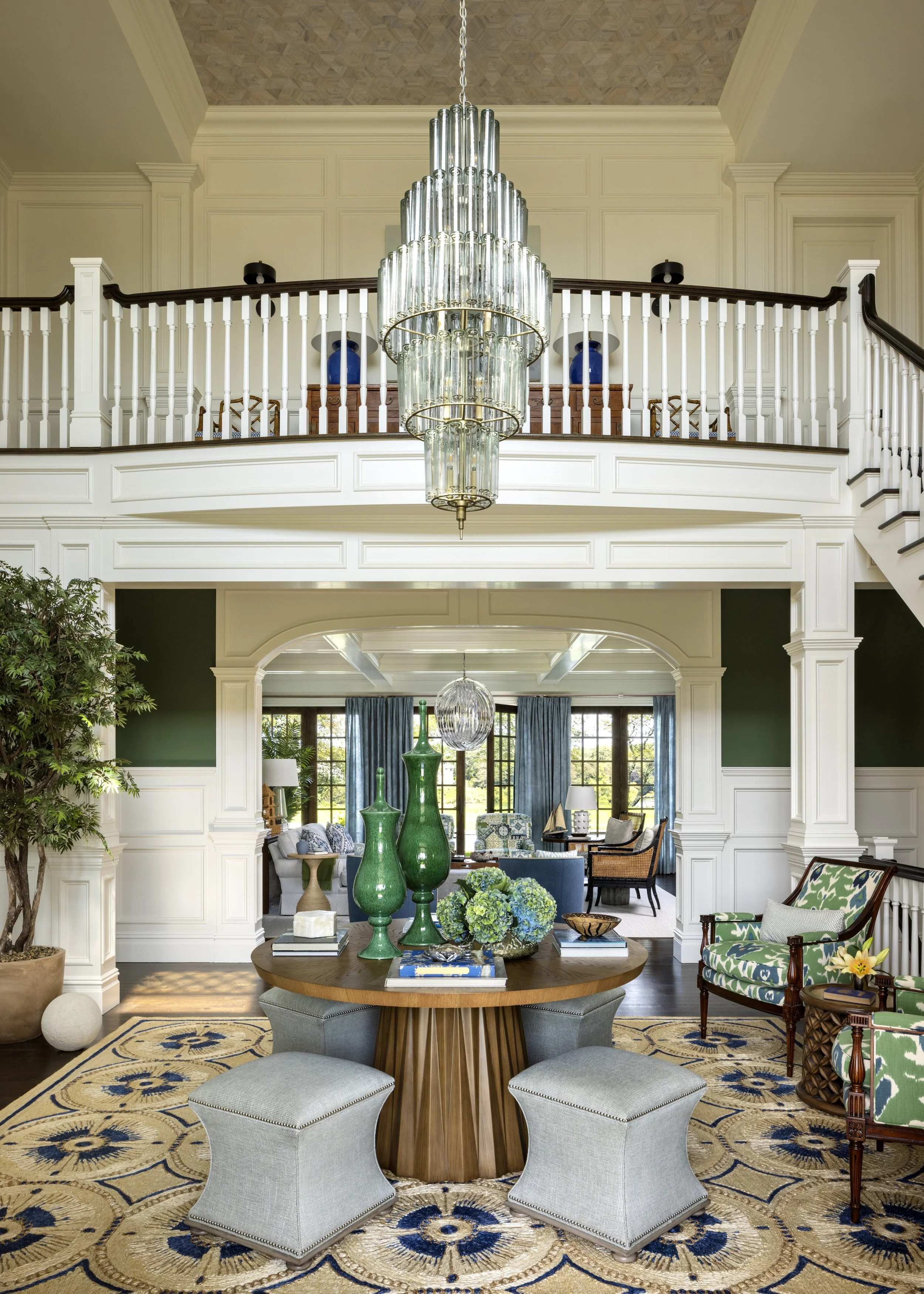 The image shows a spacious, elegant living room with a high ceiling, a large chandelier, and a balcony railing. The room features a round wooden table with green vases and floral arrangements, surrounded by various chairs and sofas with colorful cushions. The space is filled with natural light from large windows with blue curtains, and is decorated with potted plants, books, and decorative objects, combining traditional and modern styles.