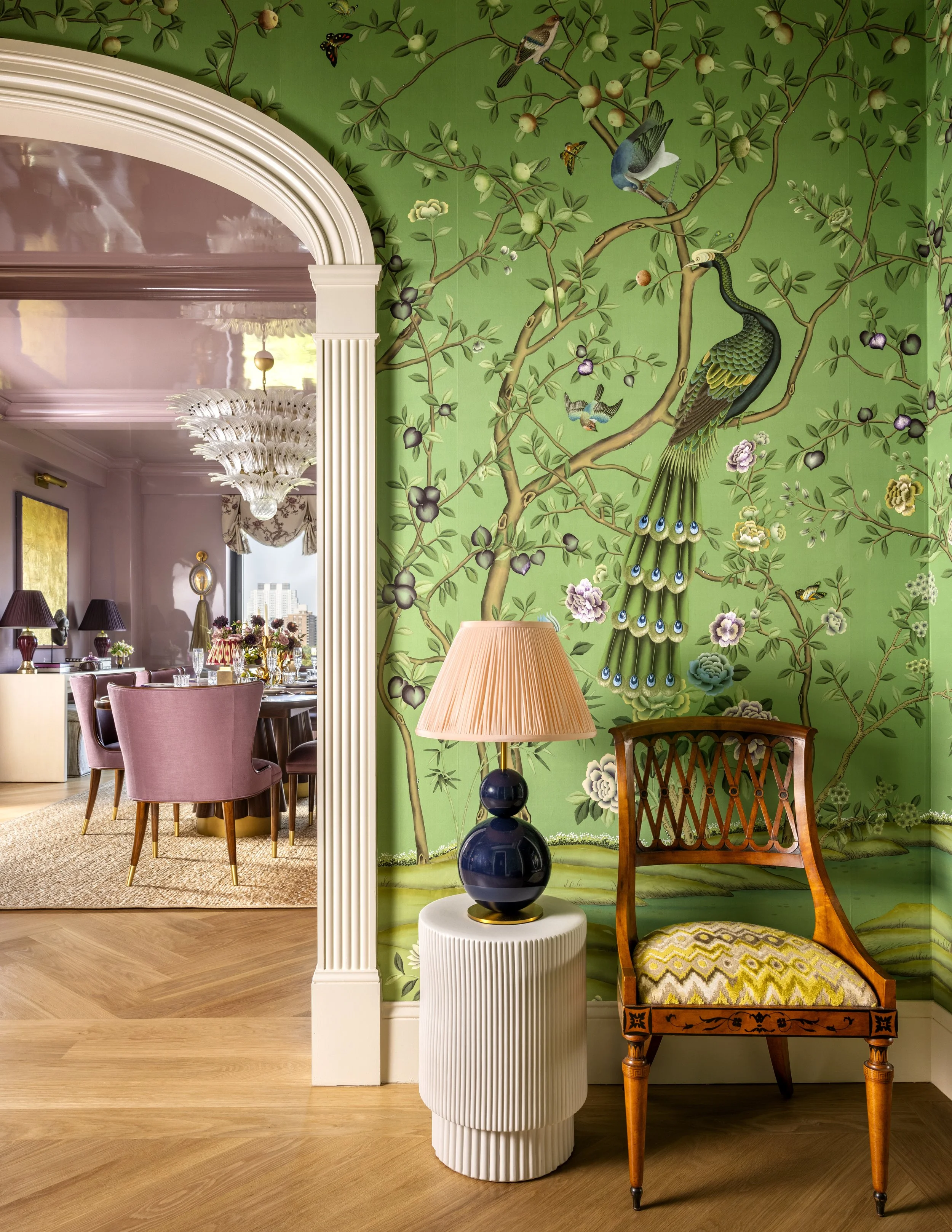 A room corner with a green wall featuring a detailed mural of a tree with birds, butterflies, and flowers. In the foreground, a wooden chair with a patterned yellow cushion and a vintage orange frame is next to a white textured side table with a dark blue and black ceramic lamp with a pink lampshade. The adjacent room visible through an archway has pink chairs, a large white chandelier, and a dining area.