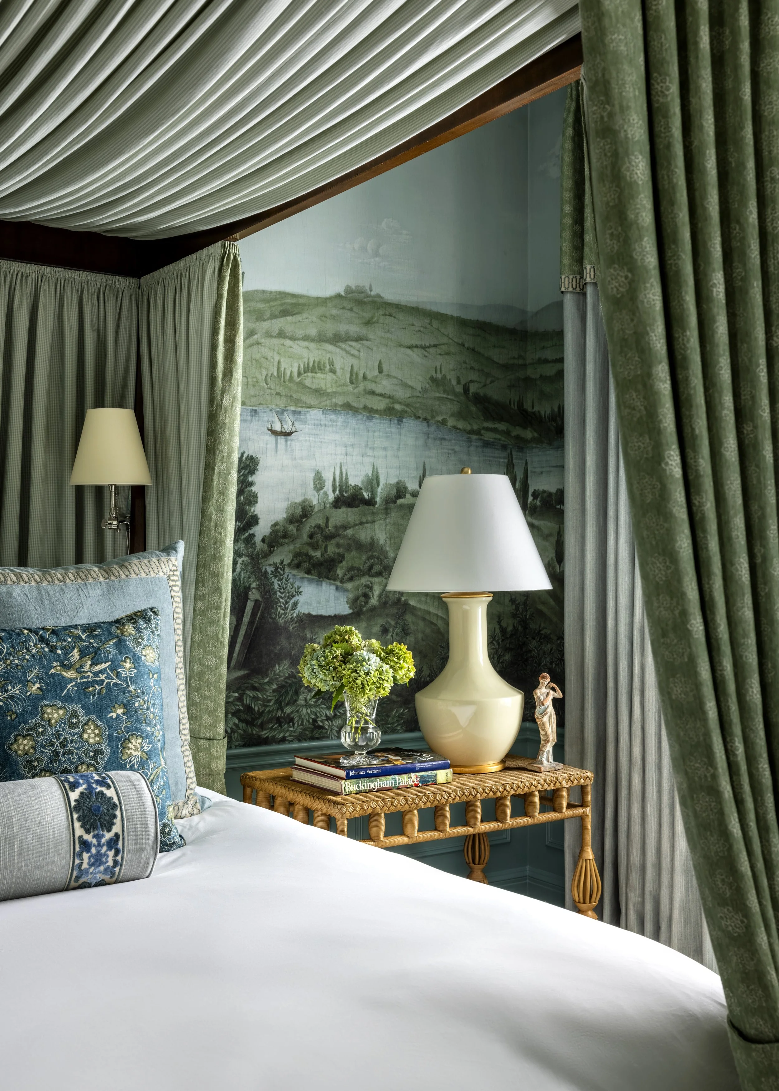 A bedroom with a white bed, decorative blue and patterned pillows, and green curtains. A wicker side table holds a stack of books, a cream-colored lamp, a small sculpture, and a vase of green flowers. A landscape mural featuring green hills, trees, a
