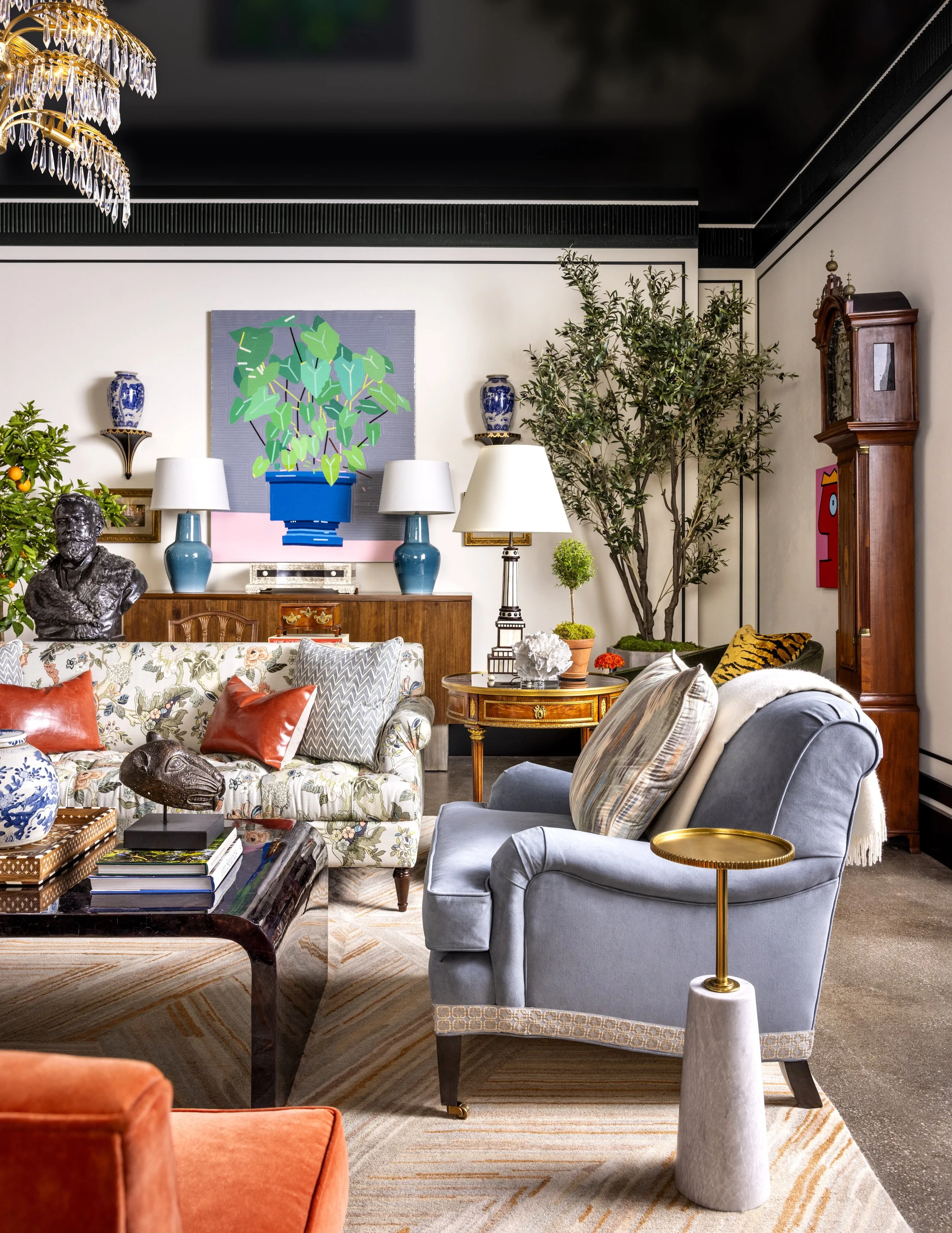Interior of a living room with a floral sofa, orange armchair, blue armchair, and side table with a gold lamp. Wall decor includes a large abstract painting of green leaves, vases, and a vintage grandfather clock. There are plants, lamps, and decorat