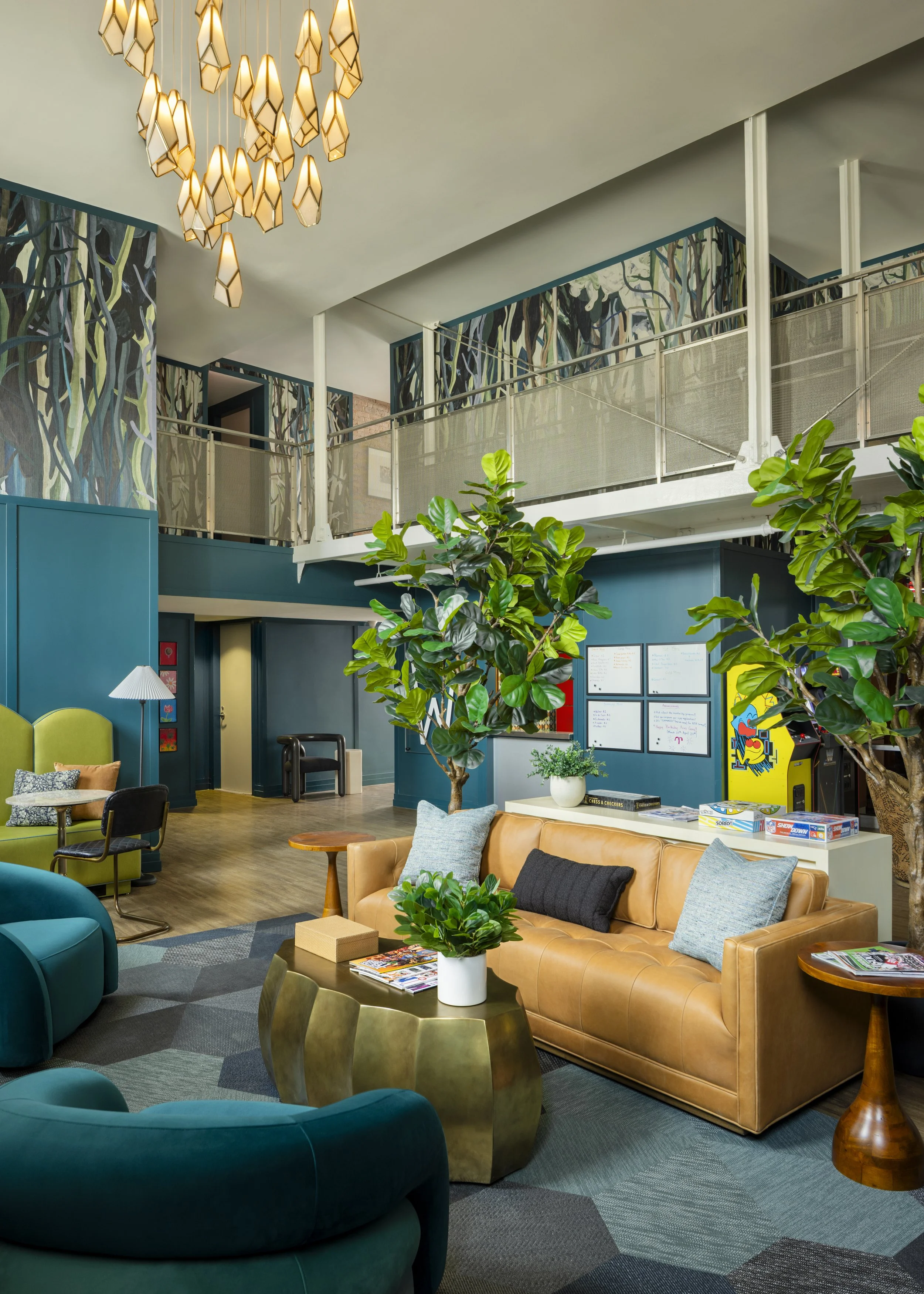 A modern, colorful lounge area with a variety of curved chairs and sofas, a coffee table with magazines and a potted plant, and a patterned wall with trees. There is a second-floor railing and a geometric chandelier hanging from the ceiling.