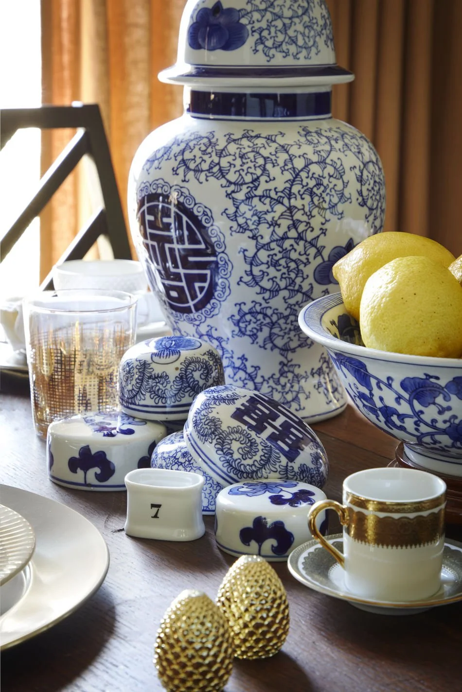 Decorative dishes and a large blue-and-white porcelain jar on a wooden table, with a bowl of lemons, a glass with a gold pattern, and golden textured eggs.