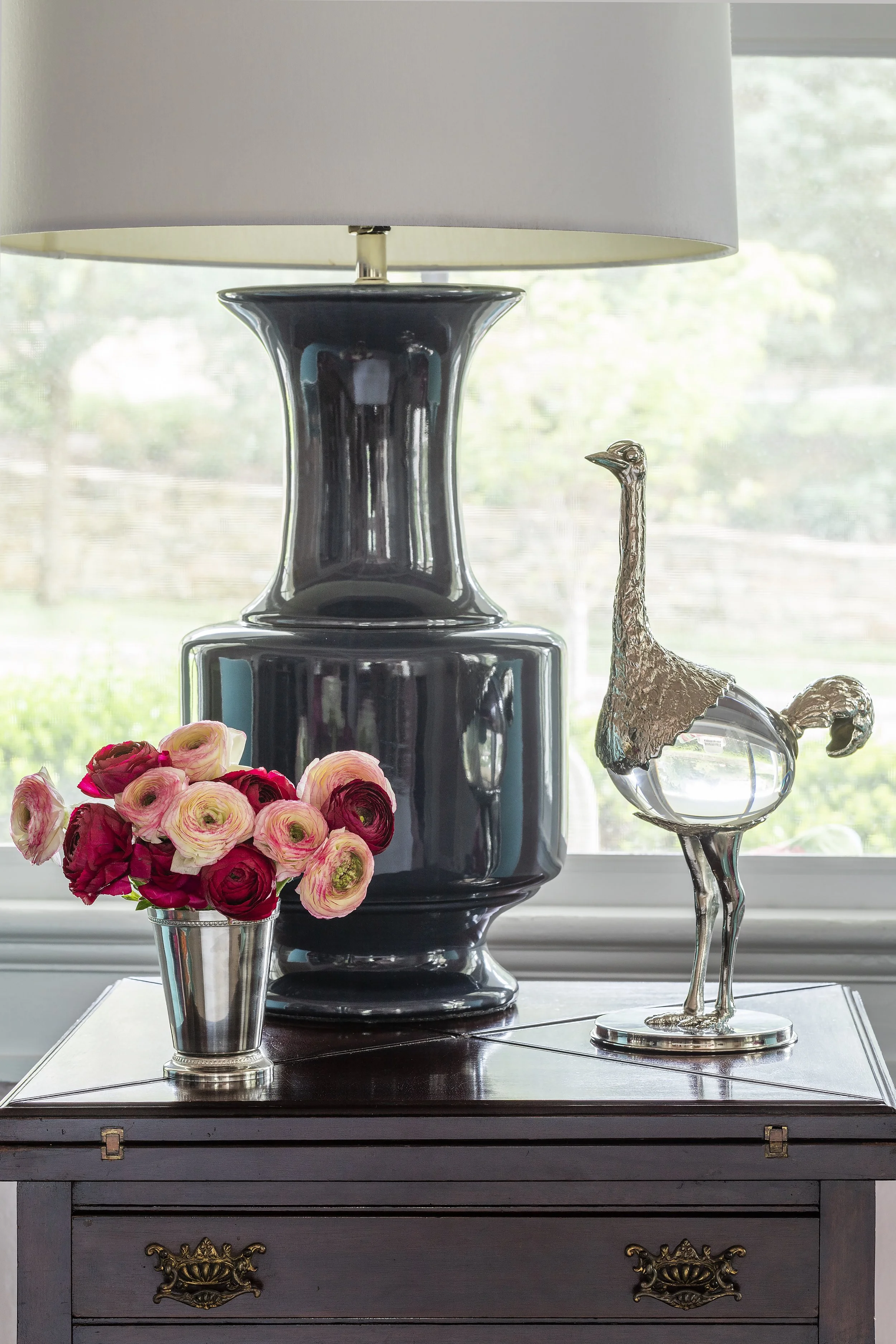 A black ceramic lamp with a white lampshade, a decorative silver flamingo figurine, and a bouquet of pink and red ranunculus flowers in a metallic vase, all on a dark wooden table against a window with a blurred outdoor view.