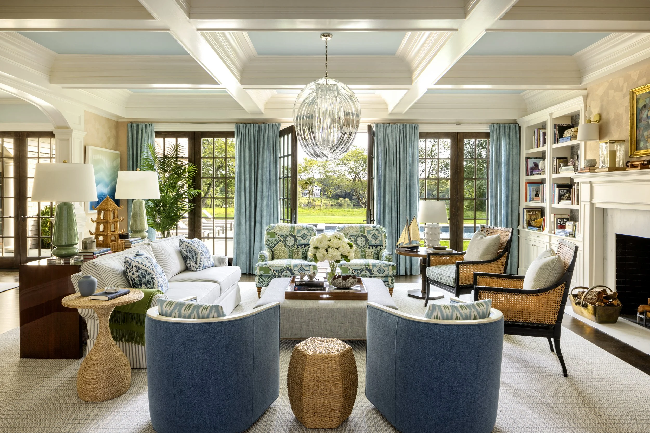 A bright, spacious living room with large windows overlooking a green landscape, featuring a white sofa, blue armchairs, a coffee table with flowers, various lamps, shelves with books, and blue curtains.