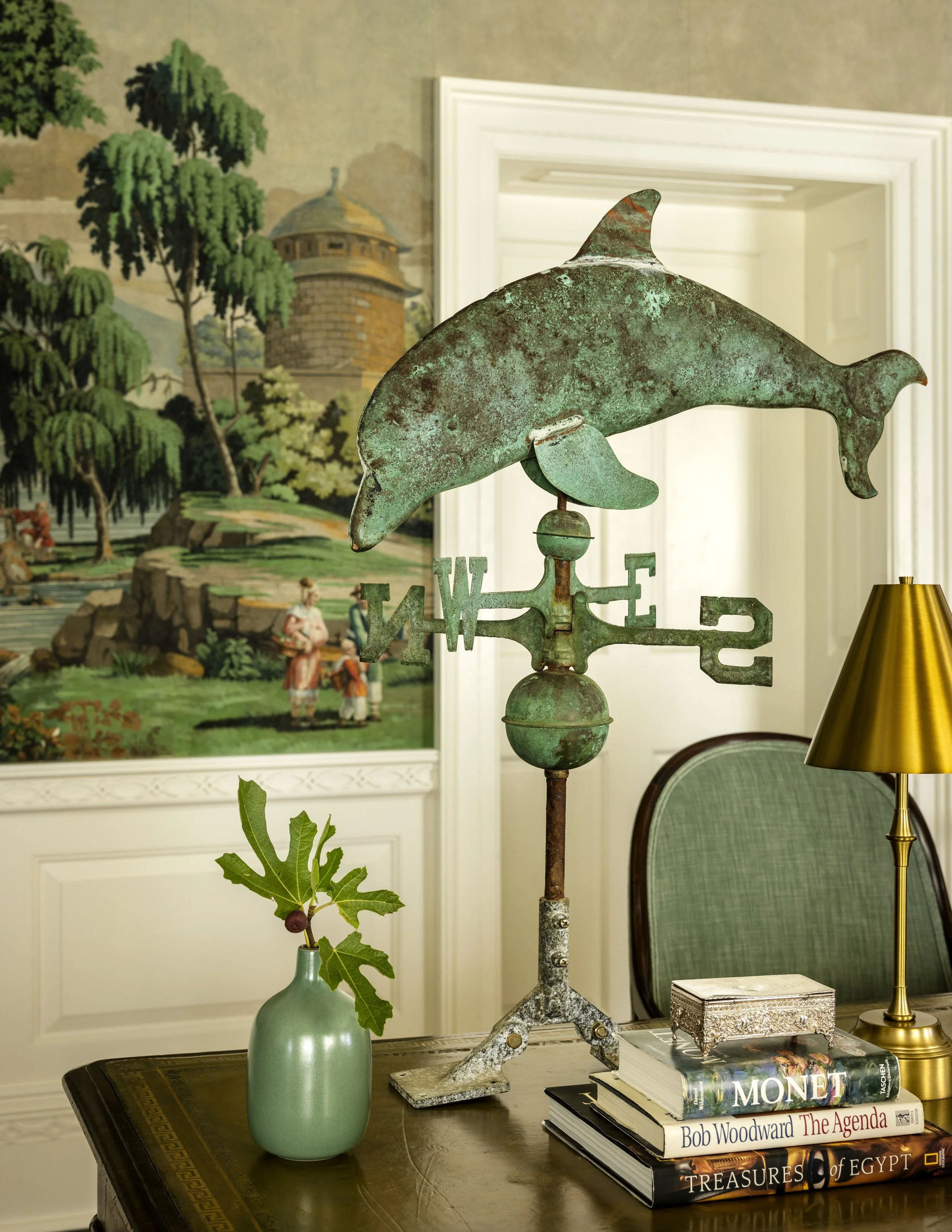 A decorative metal dolphin weather vane on a stand in a room. Behind it, there is a painting of a landscape with trees, rocks, and a tower. On the table, there is a green vase with an American oak leaf and berry branch, a stack of four books, a small