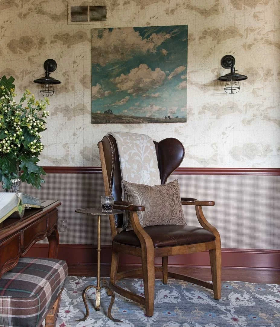 An interior scene with a brown leather armchair with a decorative white throw and a brown pillow, next to a small side table with a glass of water, a large green leafy plant with white berries on a wooden table, and a wall with a sky painting, black 