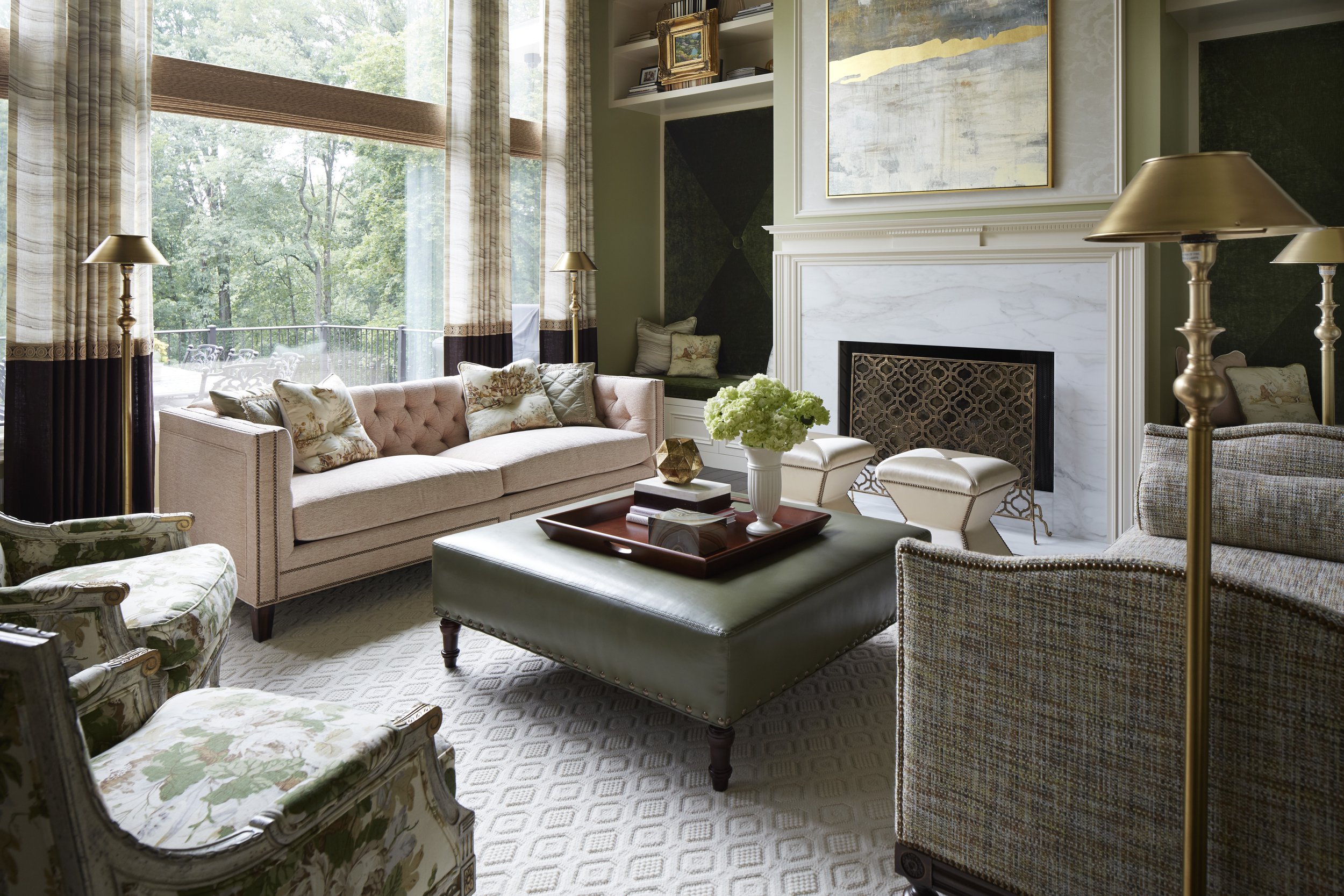 Living room with large window, beige sofa with pillows, floral armchair, patterned carpet, coffee table with flowers, fireplace, and gold lamps.