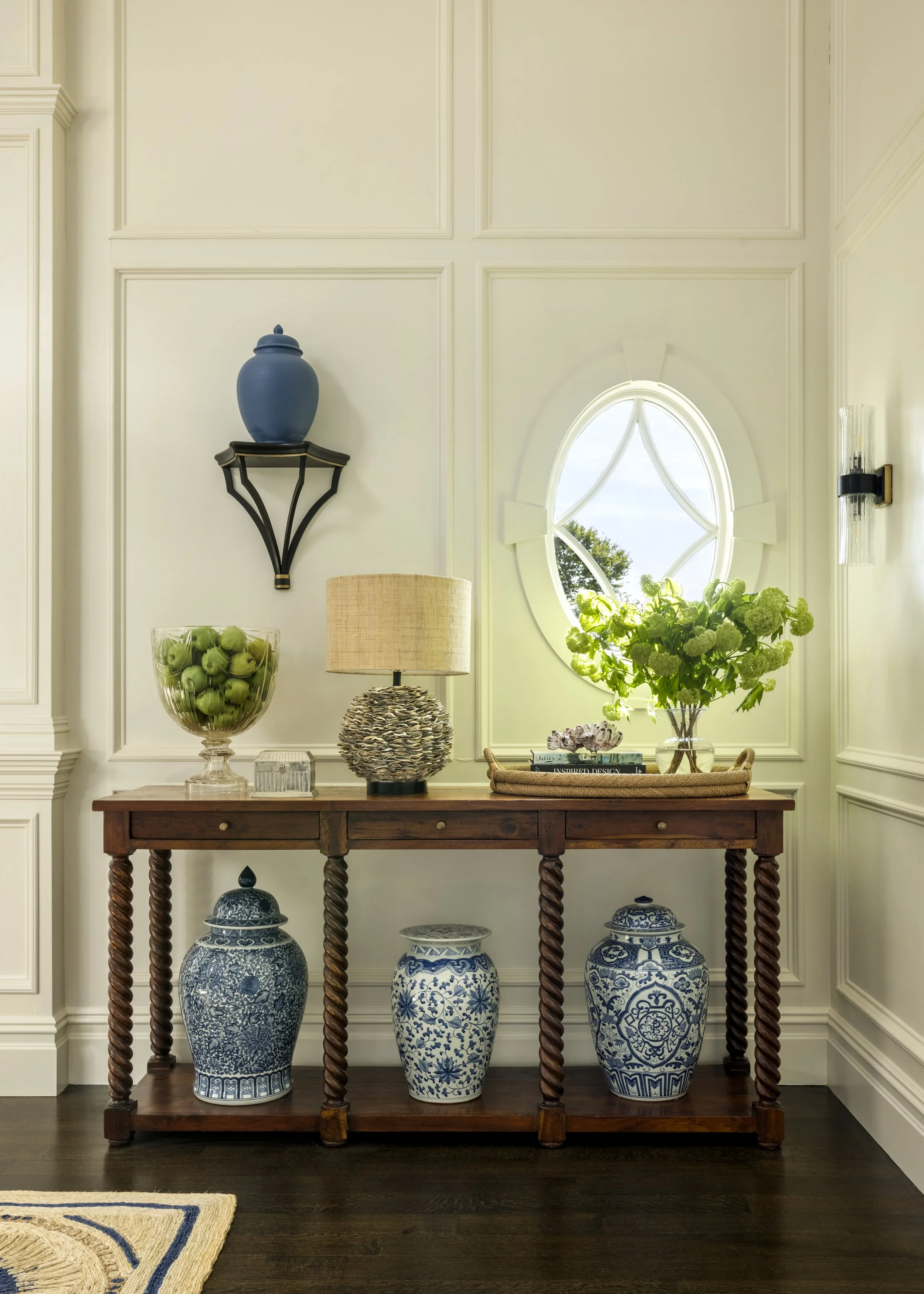 A wooden console table with twisted legs holds three large blue and white porcelain vases on a dark wood floor, with a beige and blue rug partially visible in the lower left corner; the background features cream-colored wall paneling and a small wind