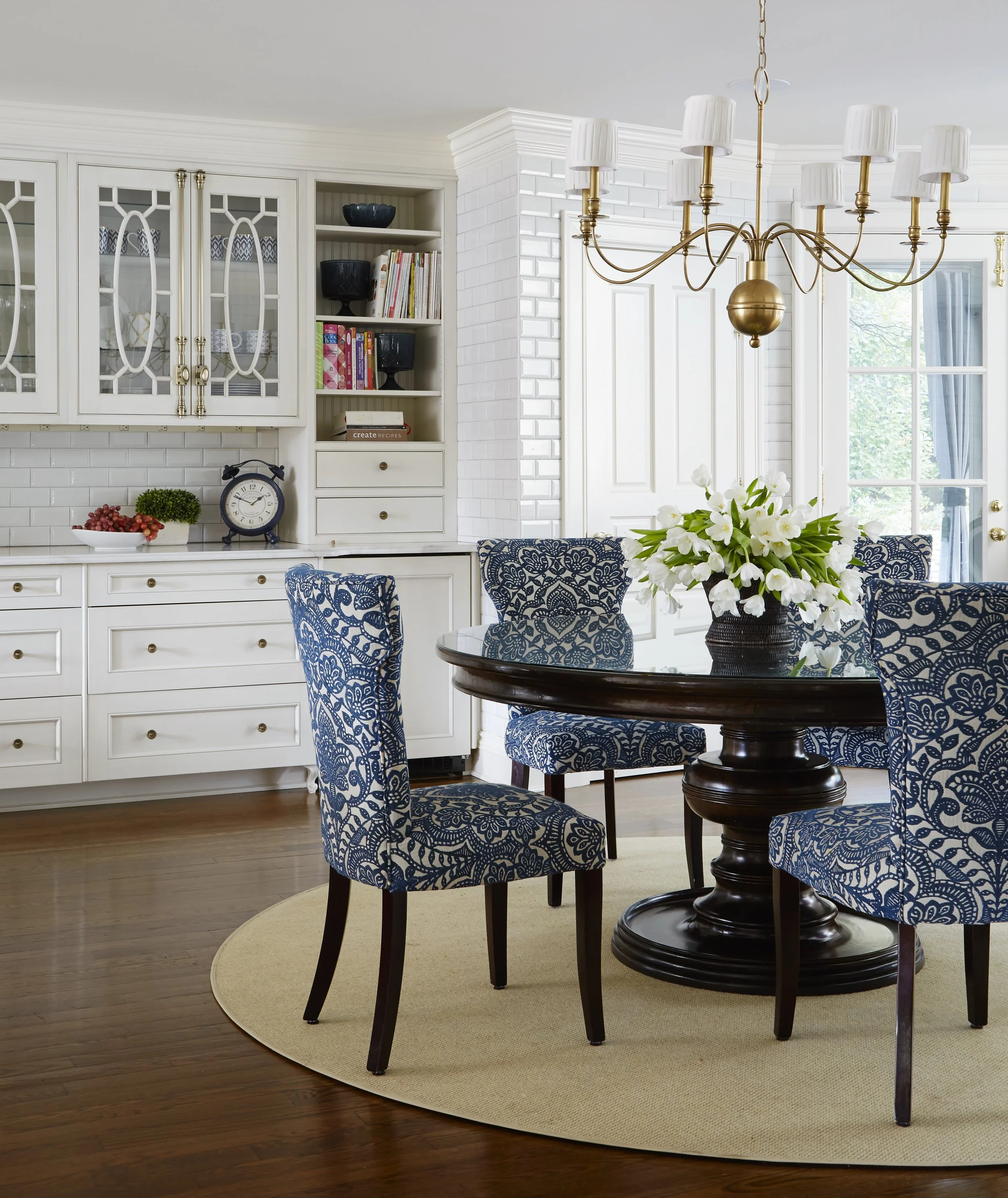 A dining room with a dark wood round table and blue patterned chairs around it. A large floral arrangement with white flowers is on the table. A beige rug is under the table and chairs. The background features a white built-in bookcase, a white paint