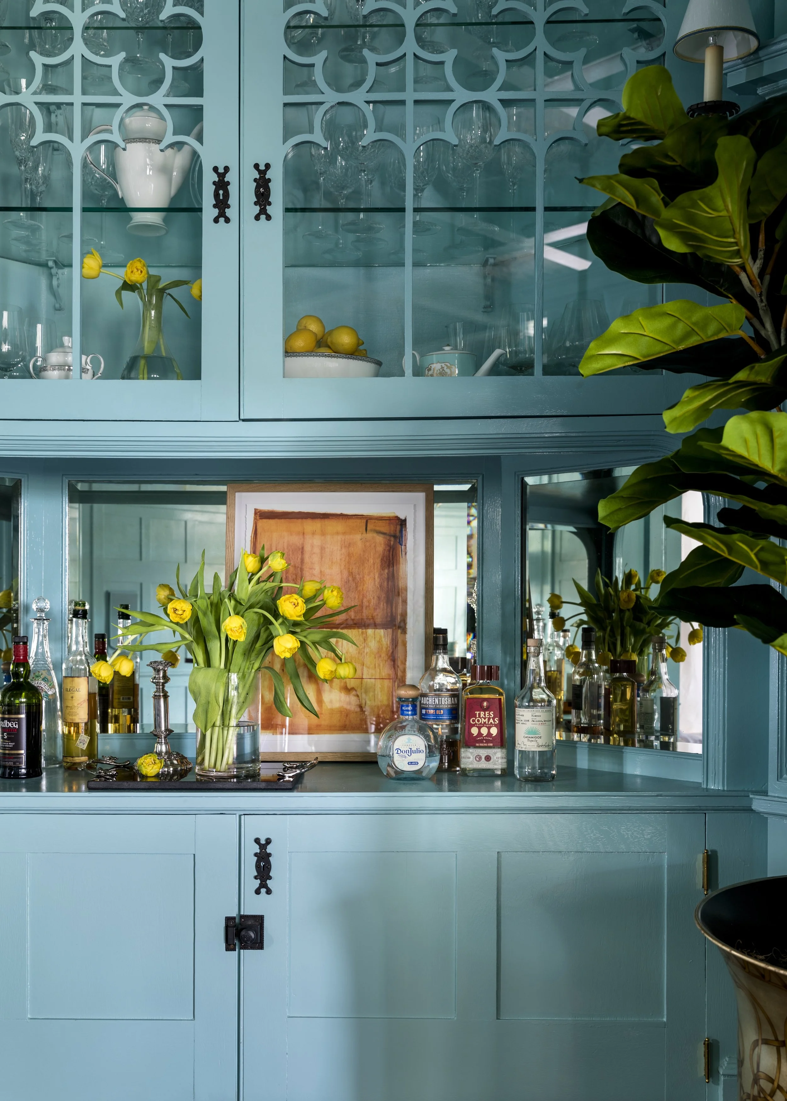 A teal-colored kitchen cabinet with glass doors displaying wine glasses and a white pitcher. Below the cabinet, a countertop features a silver tray with a large glass vase filled with yellow tulips, various bottles of alcohol, and a framed artwork. T