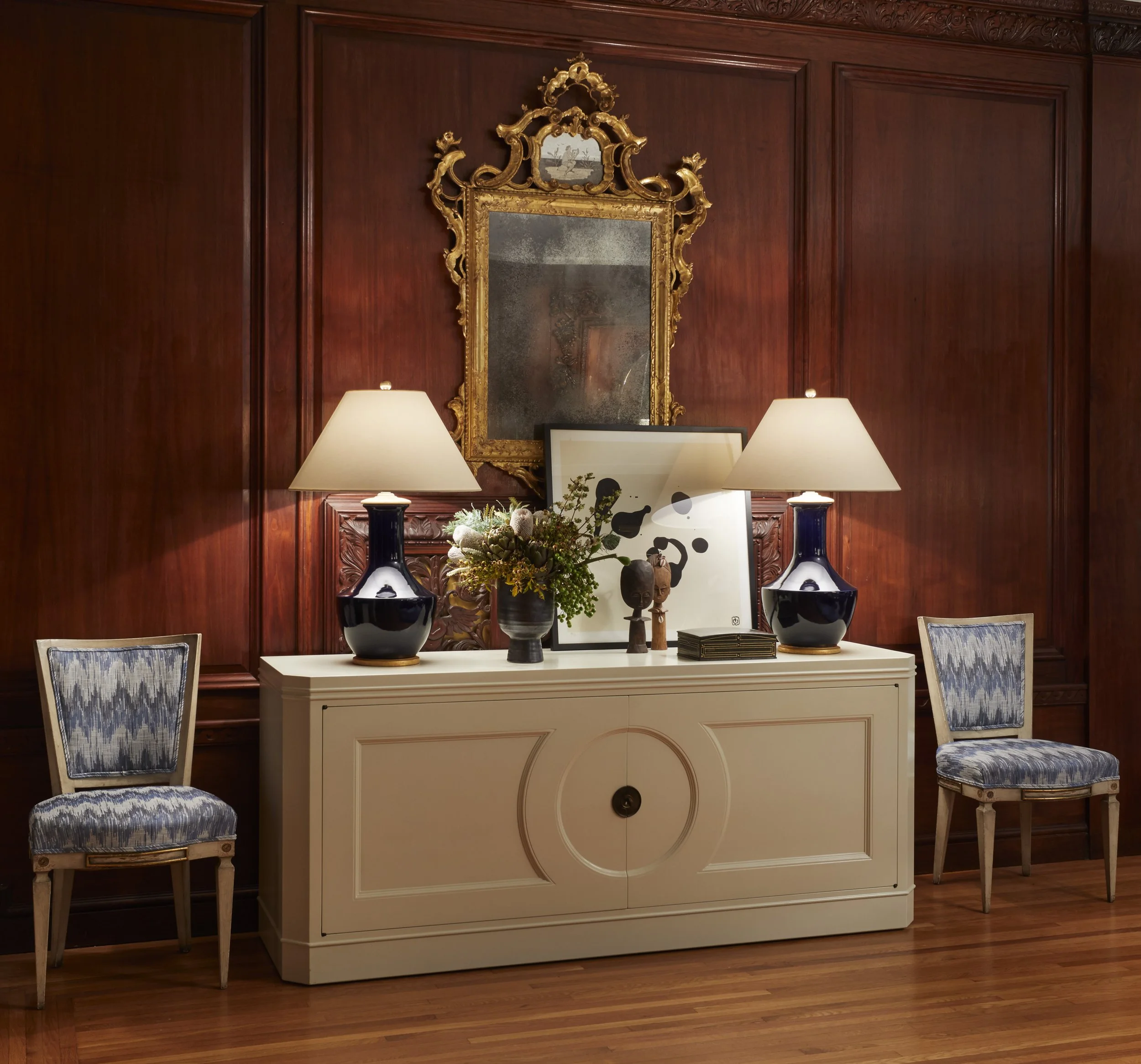 A dining room with a wooden panel wall, a white sideboard, and two blue vases with floral arrangements on top. There are two chairs with patterned cushions in front of the sideboard. Above the sideboard is a large ornate gold-framed mirror, and two t