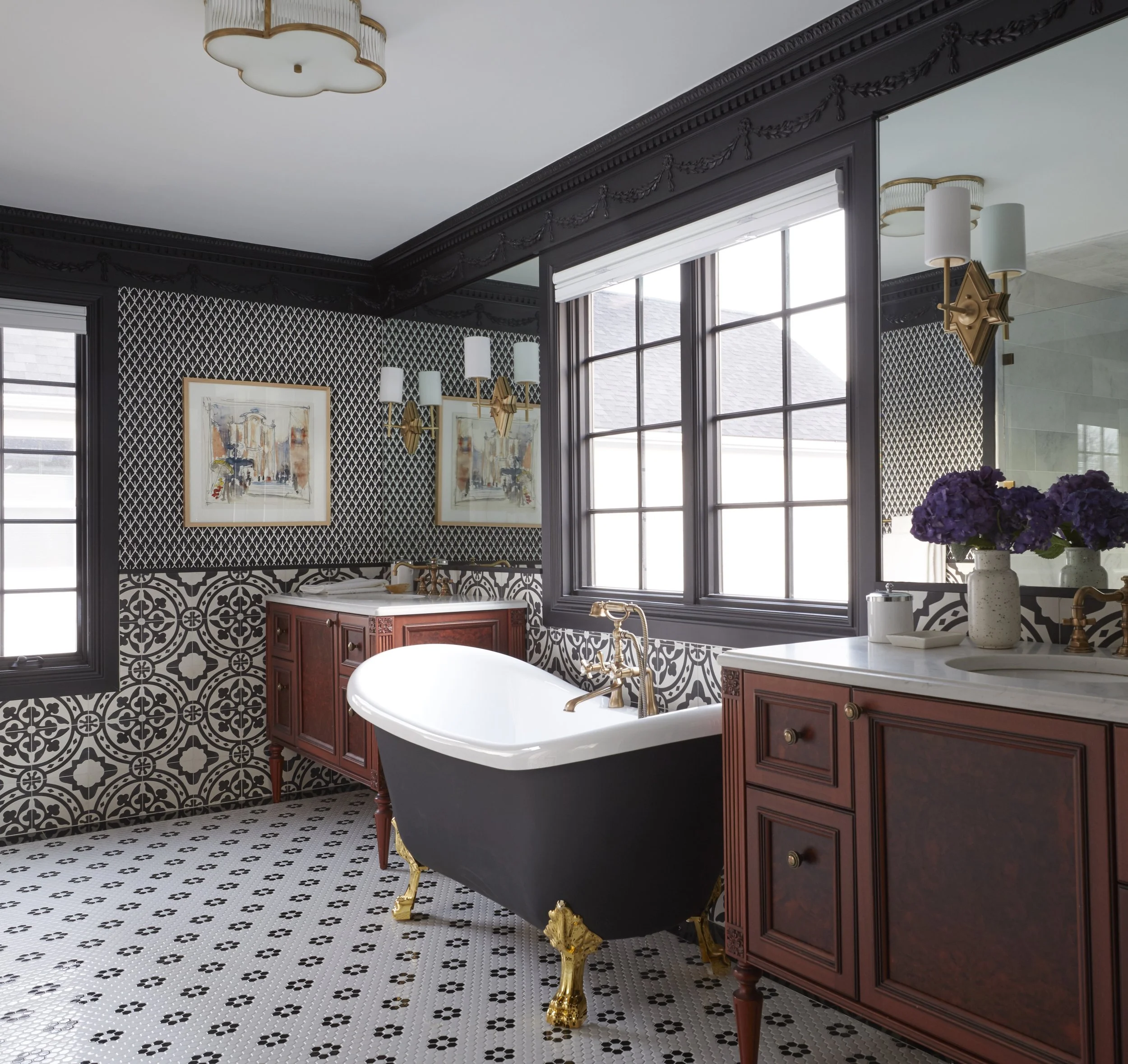 A luxurious bathroom with black and white patterned tiles on the floor and walls, a white freestanding bathtub with brass fixtures, a dark wood vanity, tall windows with white blinds, two framed paintings, and a pair of gold and white wall sconces. T