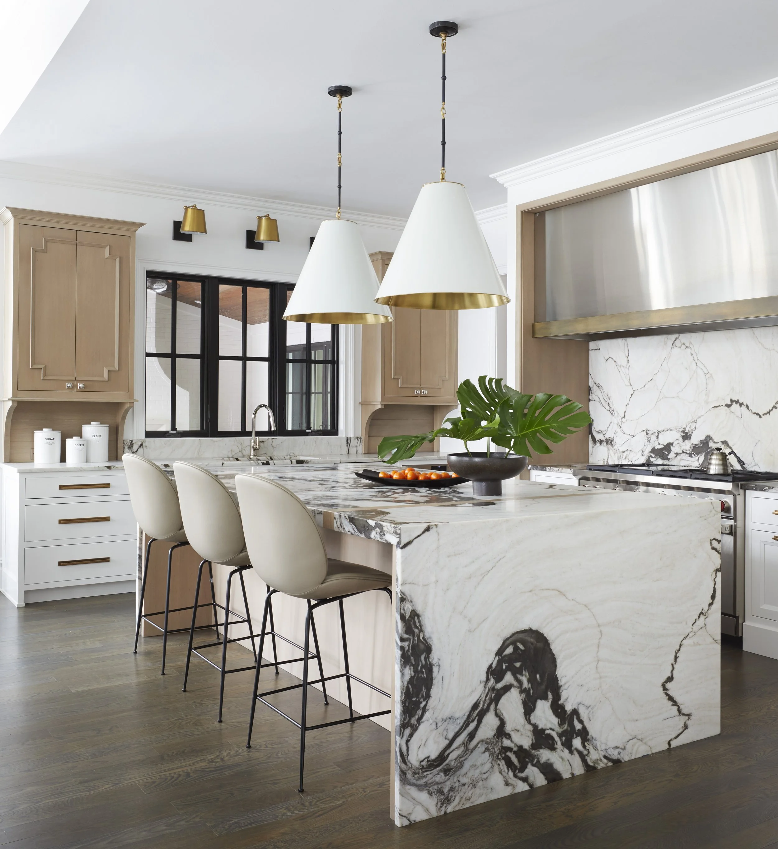 Modern kitchen interior with a white marble kitchen island featuring black and white veining. The island has a white marble top and is accompanied by four white barstools with black metal legs. On the island, there is a black bowl with a green leaf a