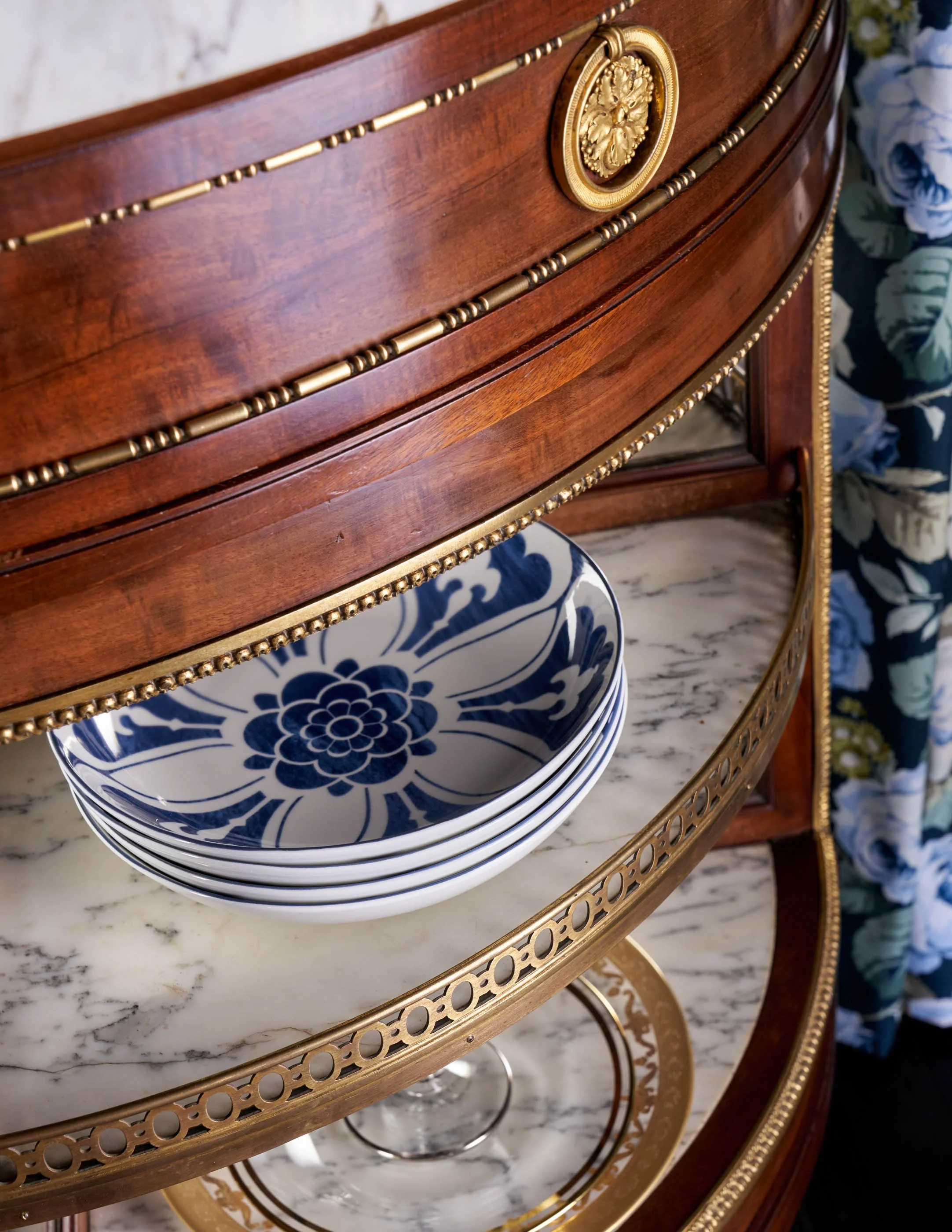 A close-up of a wooden and gold vintage side table with a marble top and a carved gold medallion, with several blue and white floral patterned plates stacked underneath.