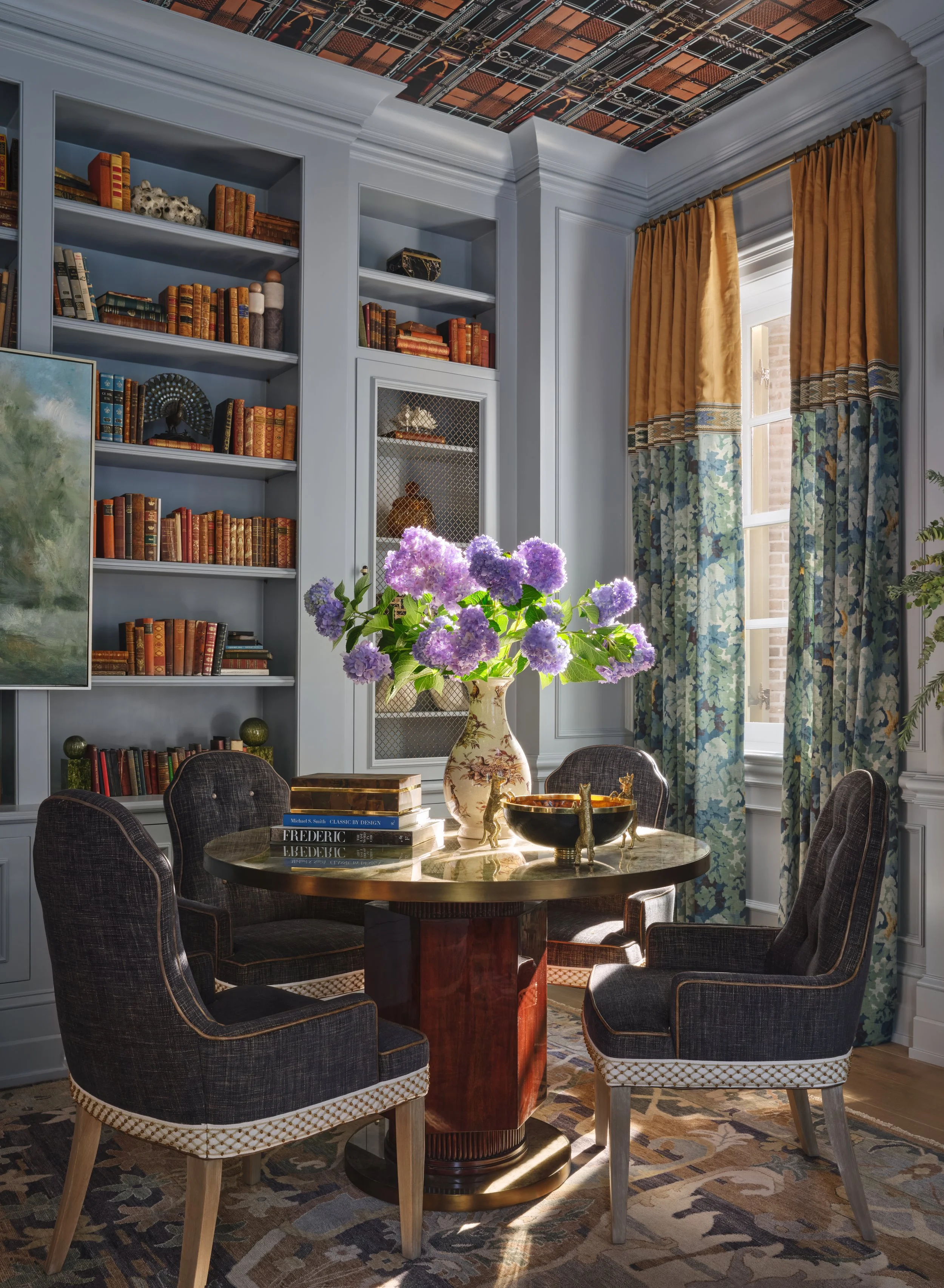 A cozy living room with built-in bookshelves filled with books and decorative items, a window with patterned curtains, a vase with purple hydrangeas on a round marble table, and four upholstered chairs around the table. The ceiling has a mosaic desig