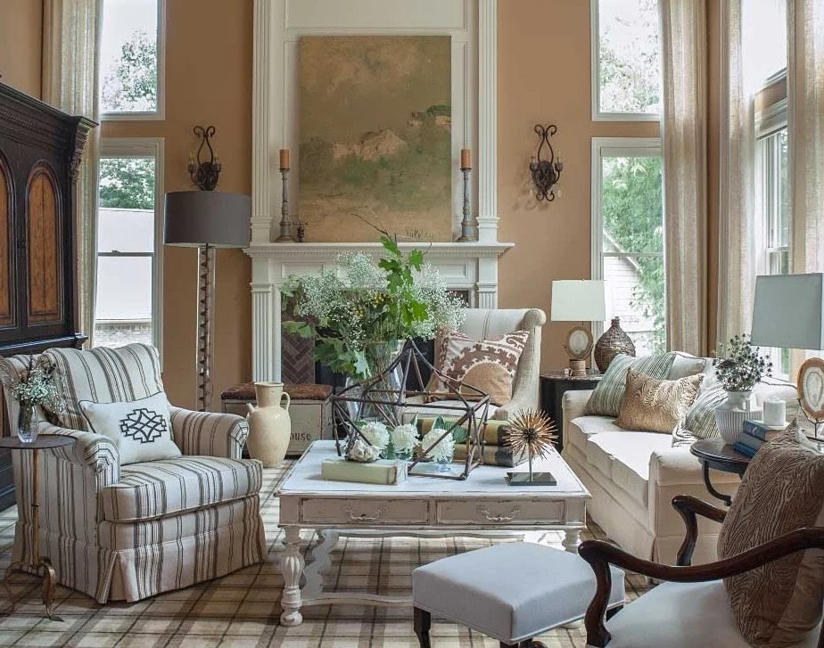 Living room with beige walls, multiple windows with curtains, a white fireplace with artwork above, and various chairs, sofas, and tables decorated with books, vases, and decorative objects.