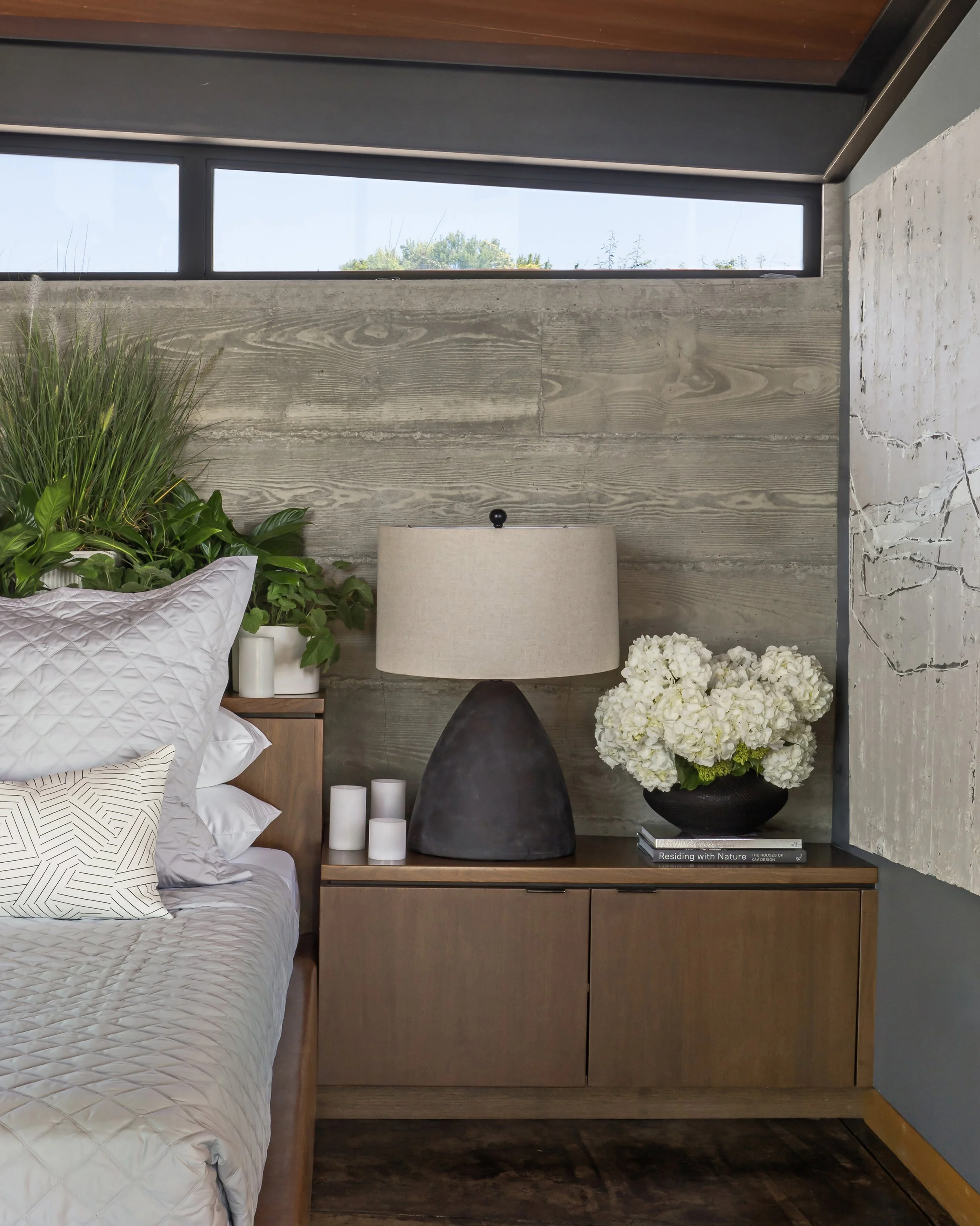 Modern bedroom with wooden wall panel behind a bedside table, decorated with a large plant in a white pot, a dark tapered lamp, three white candles, and a bouquet of white hydrangeas, with a window near the ceiling showing blue sky and green trees.