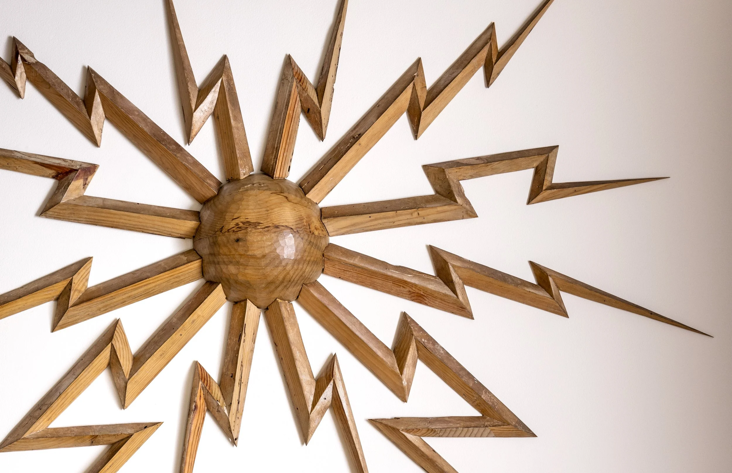 Wall decoration made of wooden pieces arranged in a sunburst pattern with a central rounded wooden piece and radiating triangular wooden segments.