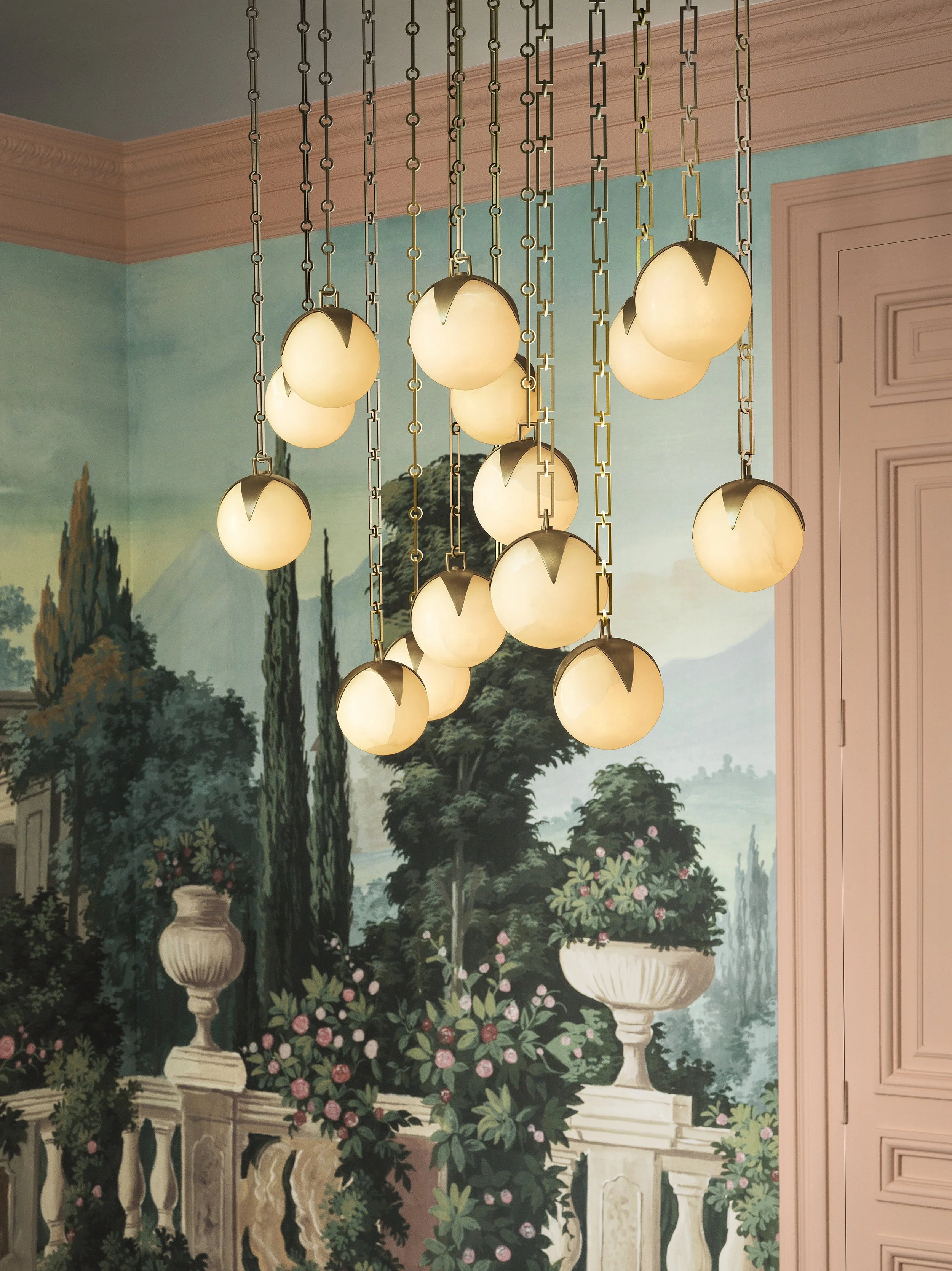 A chandelier with multiple round, frosted glass globes hanging from metal chains over a mural of a classical garden scene with trees, pink roses, and a stone balustrade.