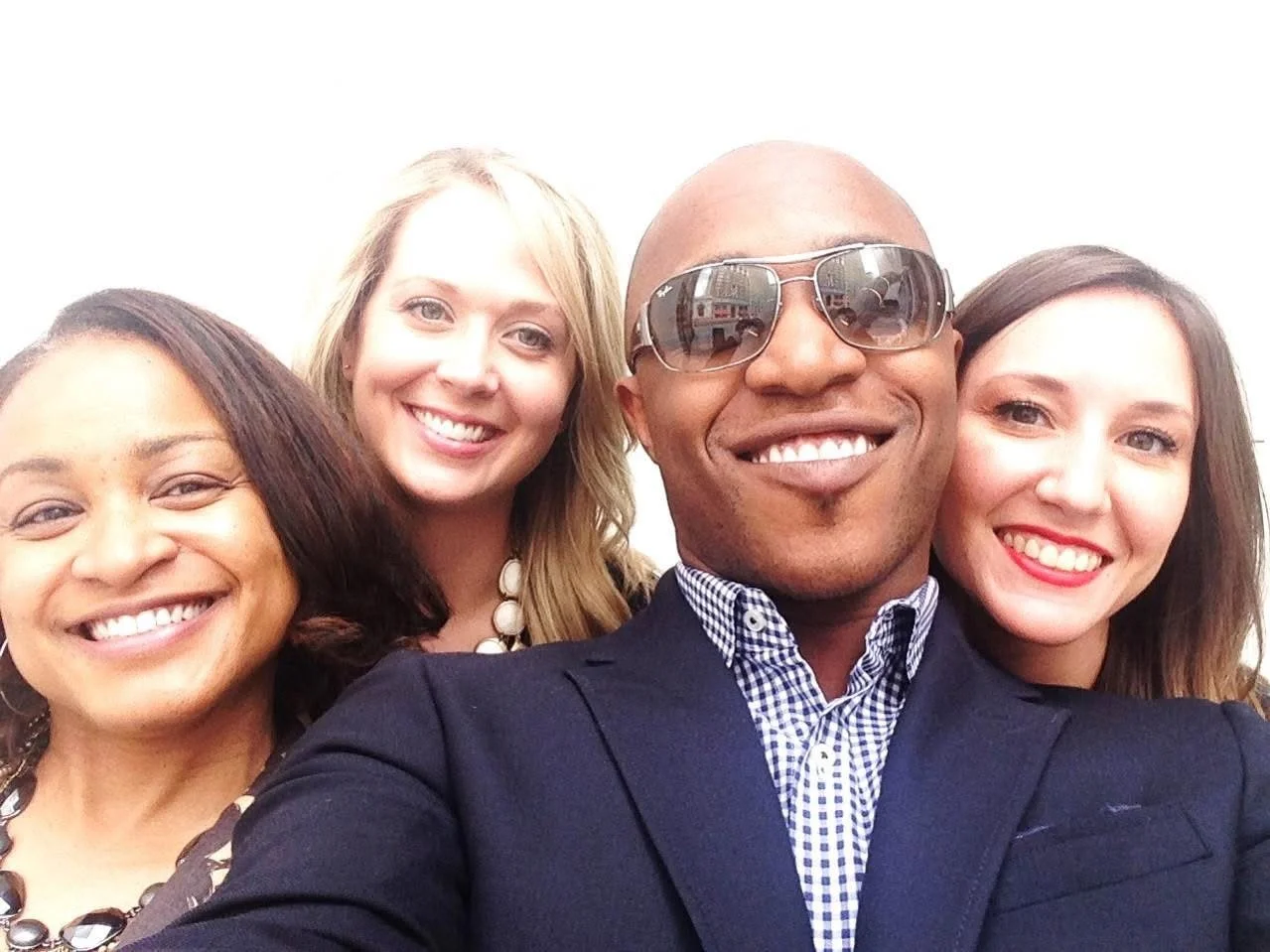 Four diverse friends smiling and taking a selfie together, dressed in business casual attire, with a white background.