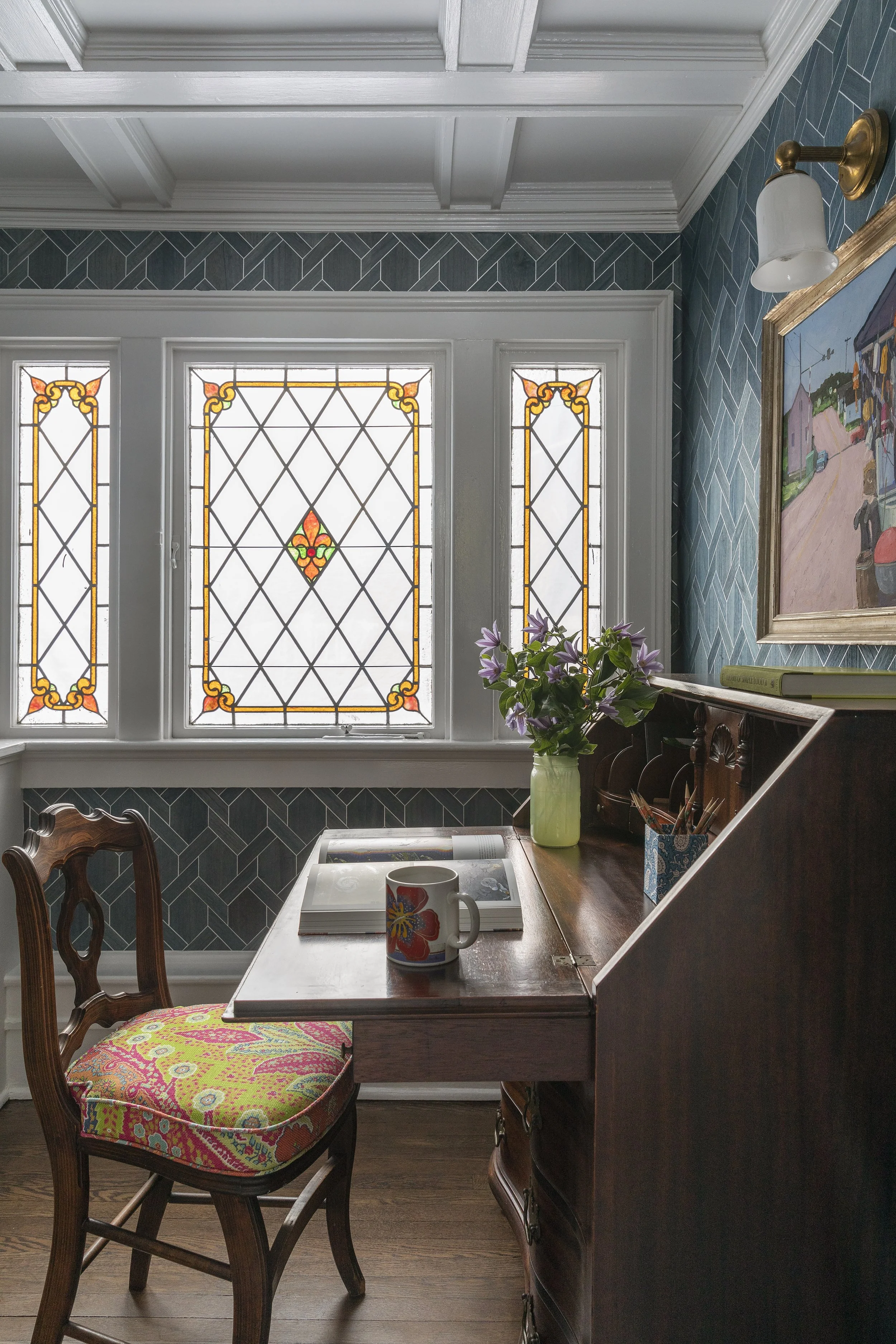 A cozy interior space featuring a wooden desk with a colorful patterned chair, a green vase with purple flowers, notebooks, a coffee mug, and a framed painting of a village scene on the wall. Behind the desk are stained glass windows with geometric a