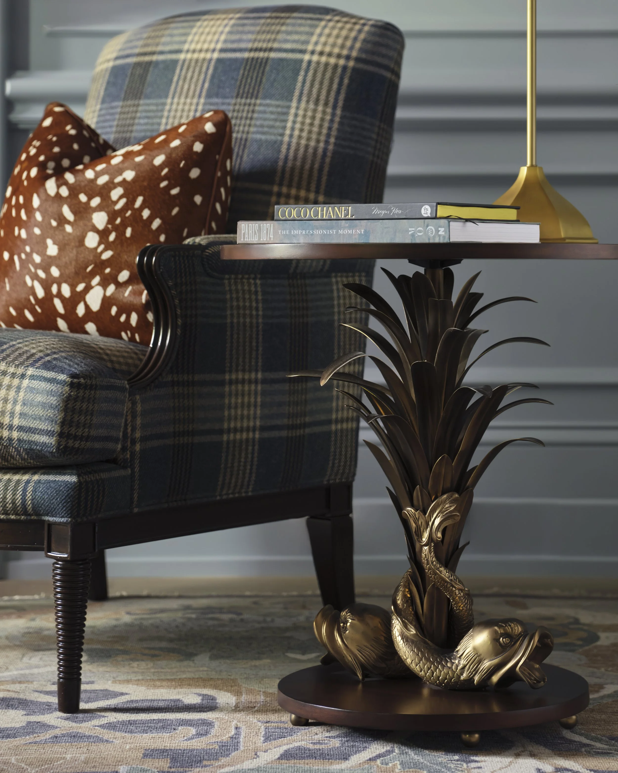 A side view of a plaid upholstered armchair with a decorative oval-shaped pillow with white spots, placed next to a round table with a gold base featuring koi fish sculptures at the bottom. The table holds three art books and a gold desk lamp. The se