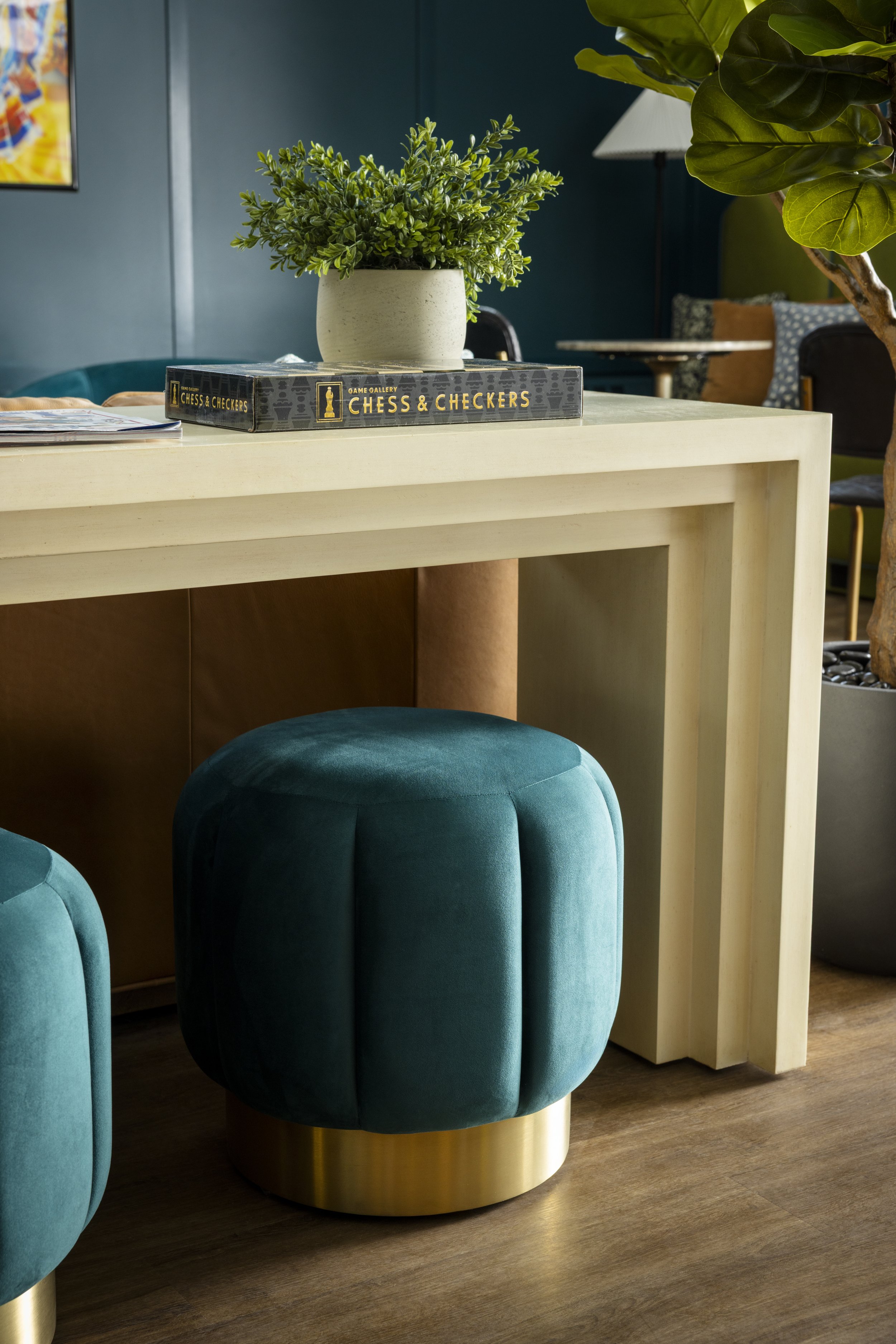 A round teal velvet ottoman with a gold base, a white table with a plant in a white pot on top, and a blue wall in the background, with a corner of a teal chair and a table with pillows visible.