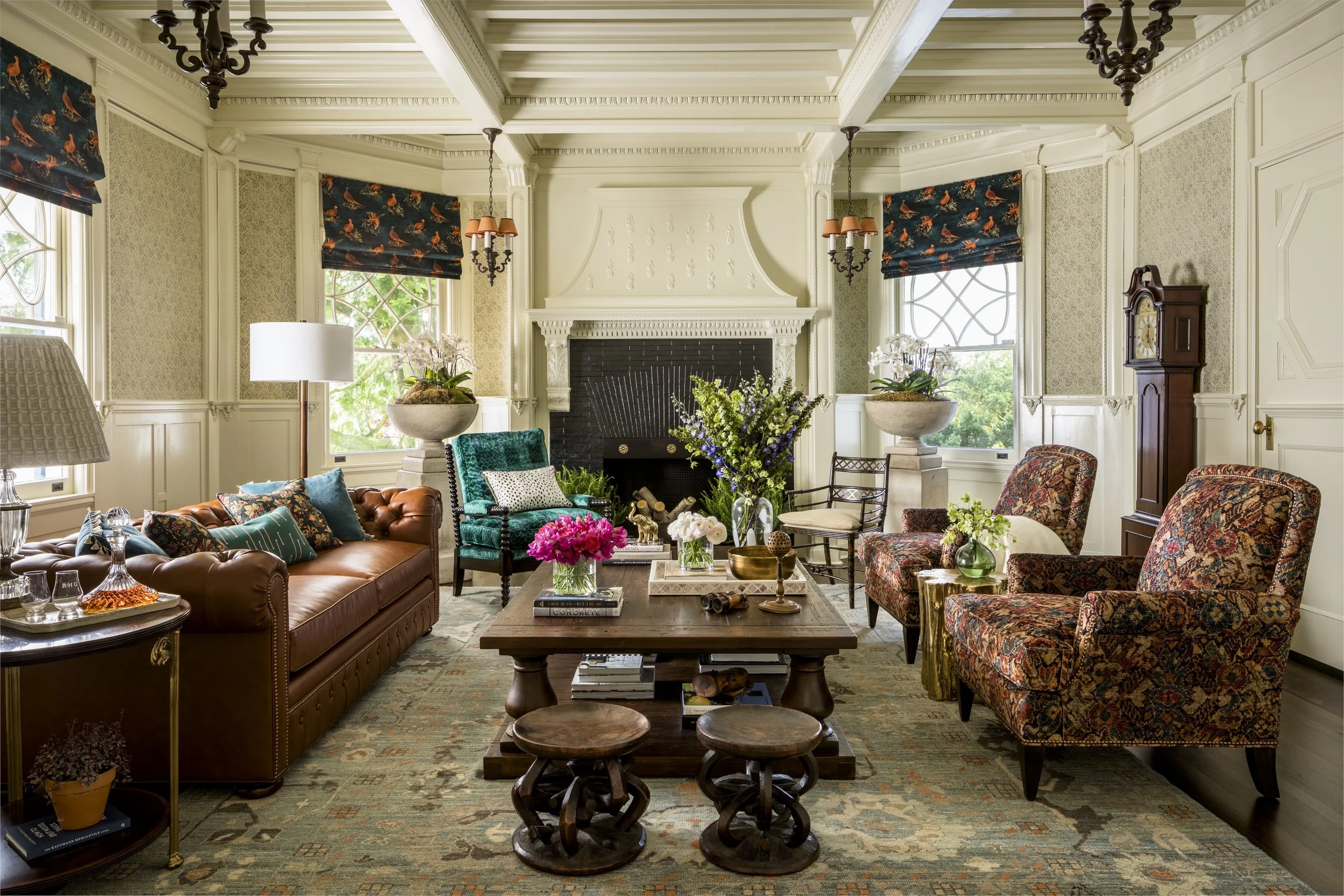 Living room with walls and ceiling painted in cream, several windows with patterned Roman shades, a fireplace, various armchairs and sofas, a wooden coffee table with decorative items, and a grand clock. The room has intricate crown molding and chand