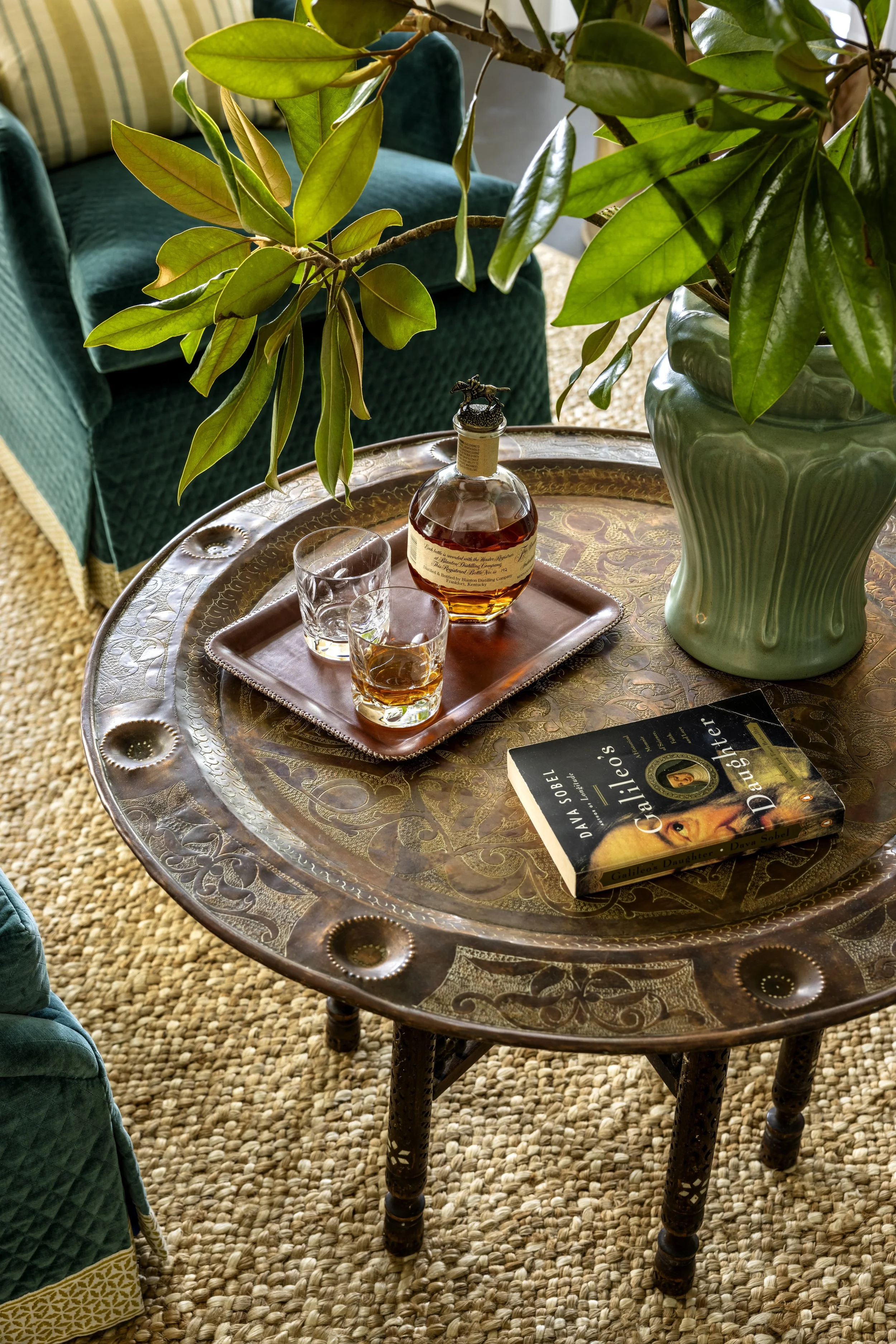 A round, carved wooden table with a book titled 'Galileo's Daughter' resting on it, a tray with two glasses and a bottle of liquor, two potted plants, and a teal chair in the background with a striped pillow.