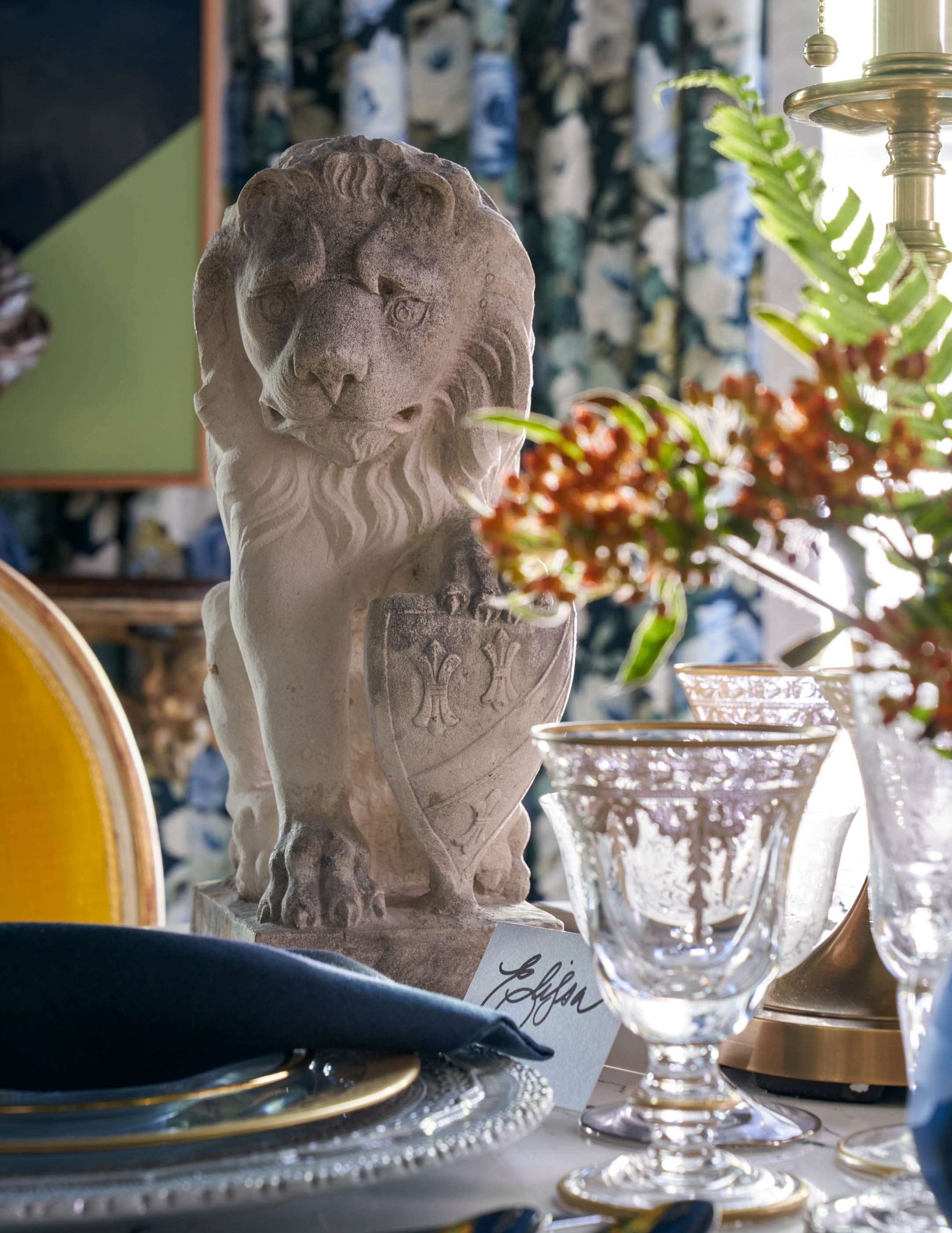 A close-up of a stone lion sculpture surrounded by glassware and decorative items on a display table, with a background of patterned fabric and sunlight streaming in from a window.