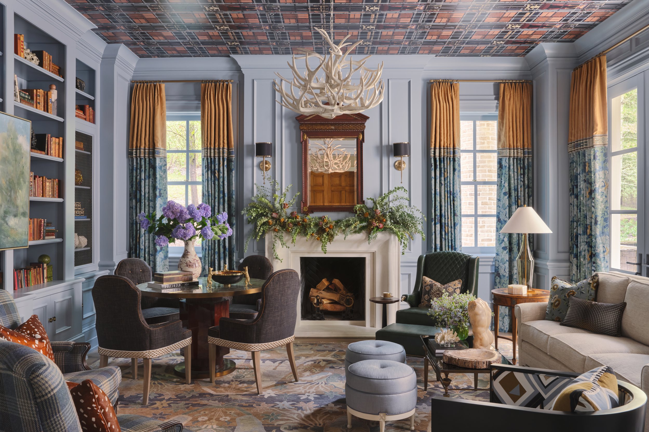 A stylish living room featuring a blue wall with a fireplace, a mirror, framed pictures, and elegant curtains. The ceiling has a patterned design, and there is a large antler chandelier hanging above. The room is decorated with colorful flowers, book