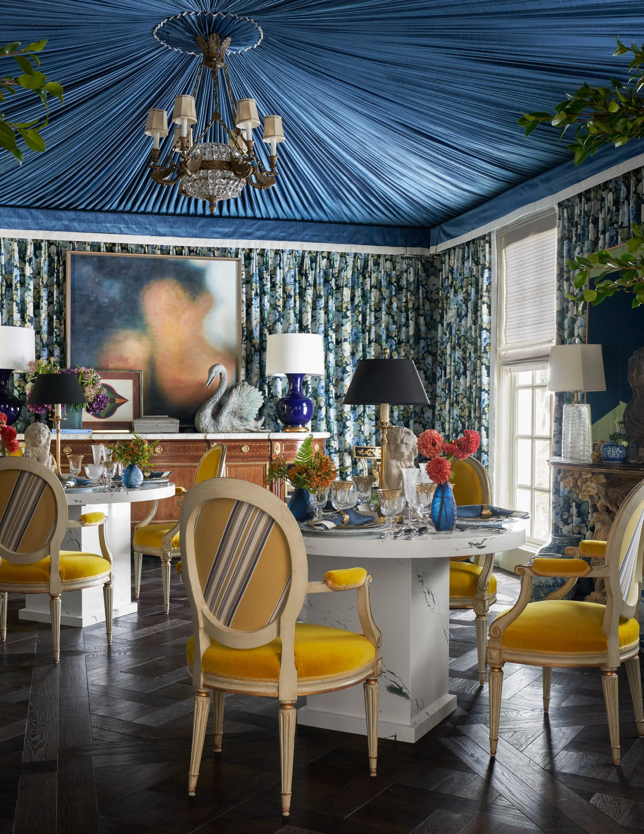 Elegant dining room with yellow velvet chairs around a white marble table, decorated with blue vases, floral arrangements, and crystal glassware. The room has floral-patterned curtains, a large abstract painting, and a blue fabric draped ceiling with