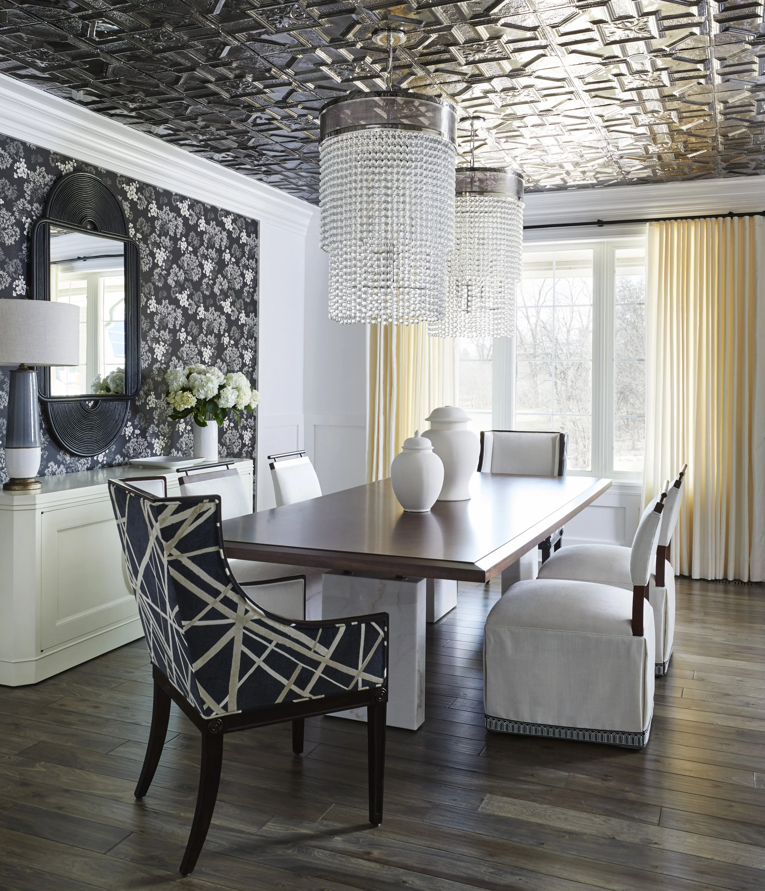 A dining room with a wooden table surrounded by white upholstered chairs, black patterned armchair, and decorative vases on the table, with large windows and cream curtains, black floral wallpaper on one wall, and a ceiling with metallic square tiles
