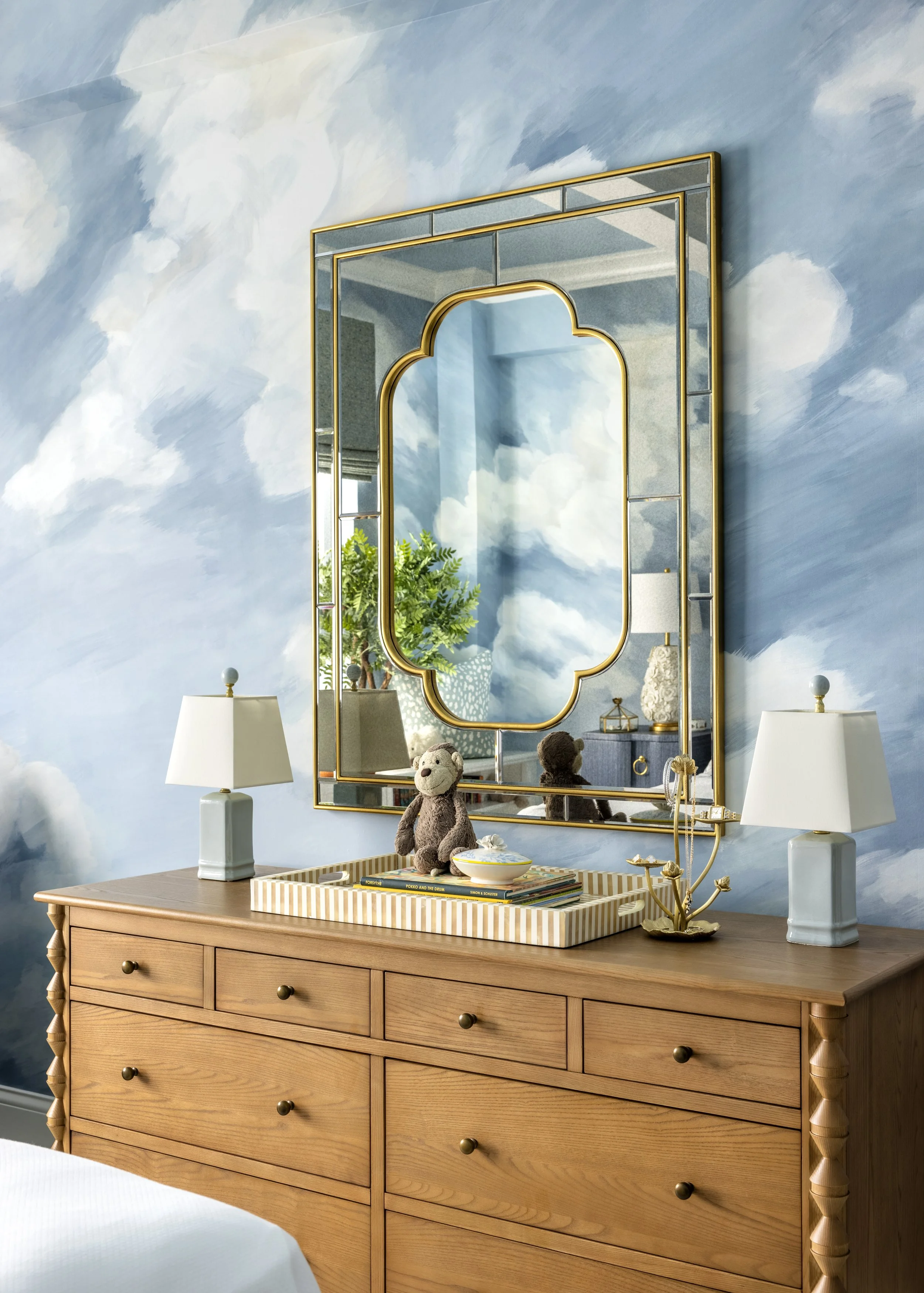 A wooden dresser with six drawers, a tray on top with books, a bowl, and stuffed animals. Two identical white lamps with rounded bases sit on either side of the tray. A mirror with gold trimming is mounted on the wall behind the dresser, reflecting g