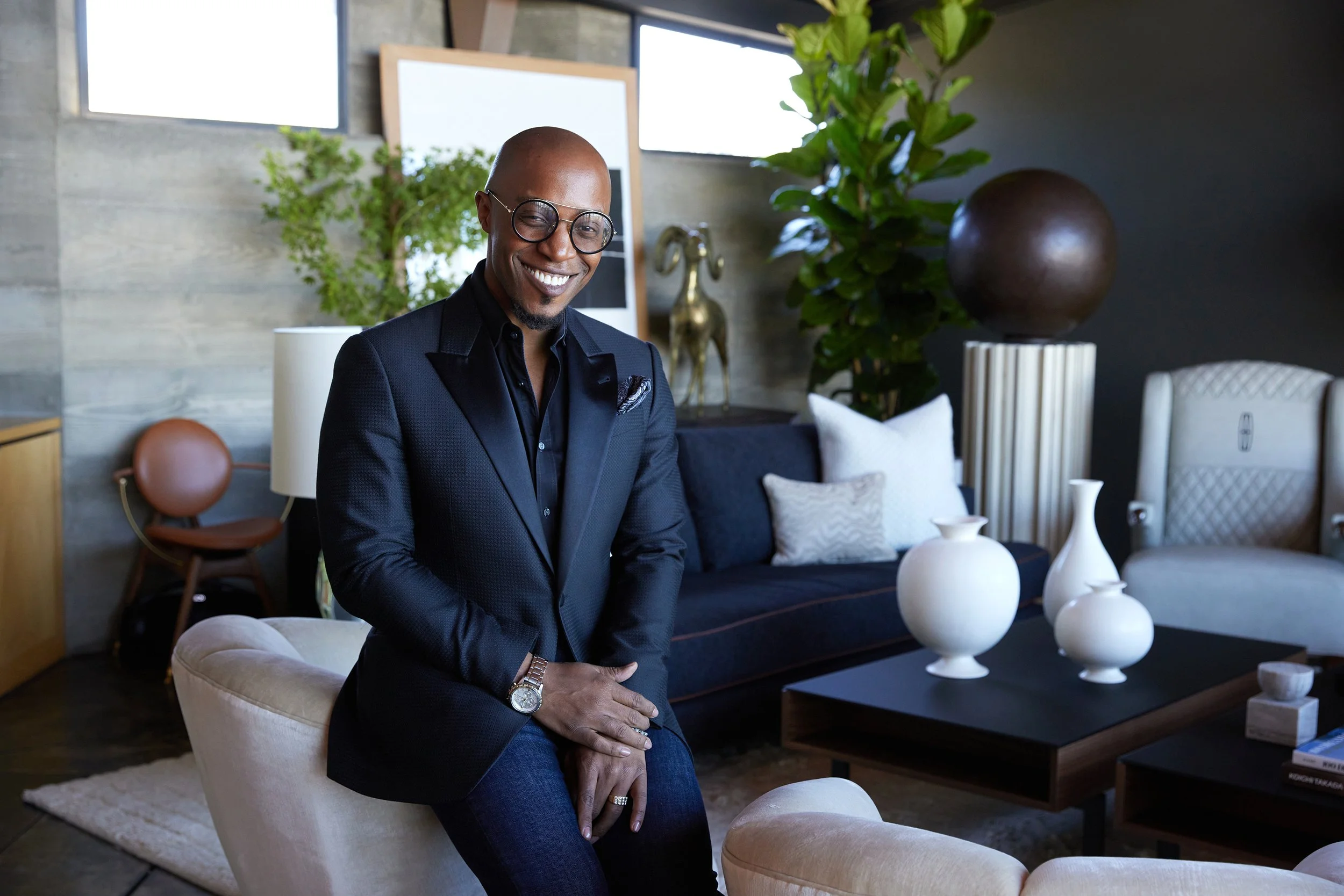 Corey Damen Jenkins, wearing glasses, a black blazer, and a watch, is sitting on a beige chair in a modern living room with decorative items and green plants.