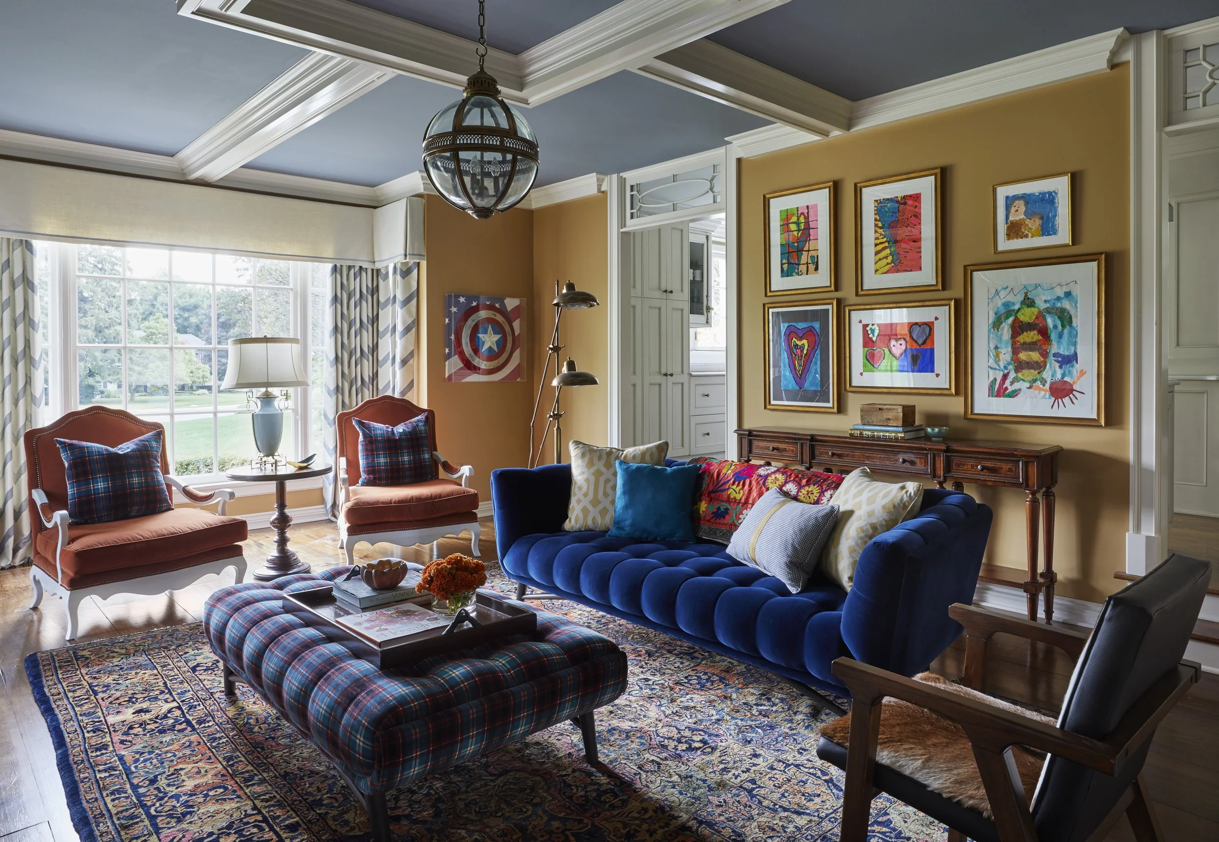 A cozy living room with colorful furniture, including a royal blue velvet couch, two peach armchairs with plaid pillows, a plaid ottoman, a patterned area rug, and various throw pillows. The room has large windows with patterned curtains, and the wal