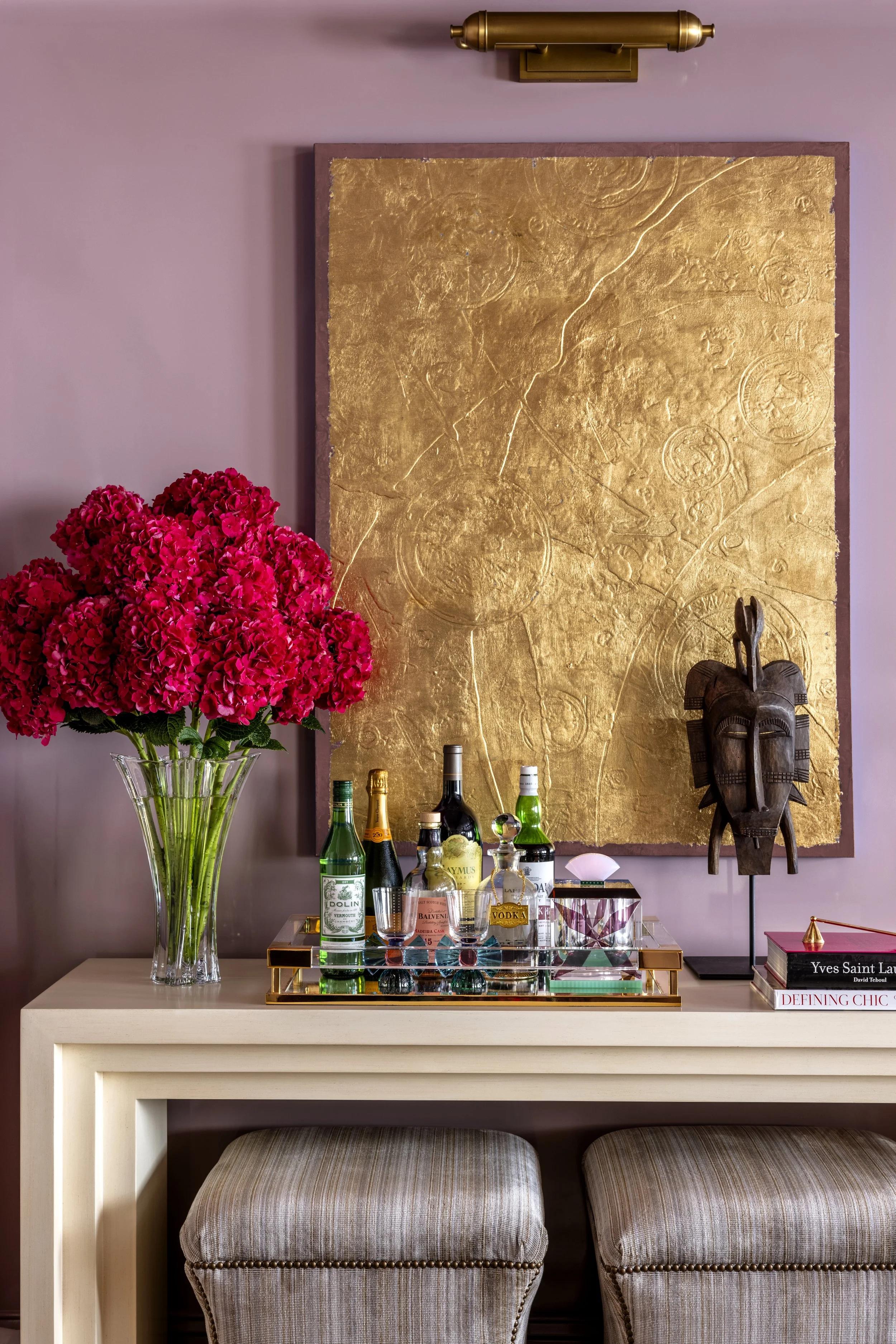 A bar setup with a glass vase of bright pink hydrangeas, a gold tray with liquor bottles and glasses, a gold abstract art piece on a mauve wall, a wooden African tribal mask, and a stack of books. Two striped upholstered stools are under a white tabl