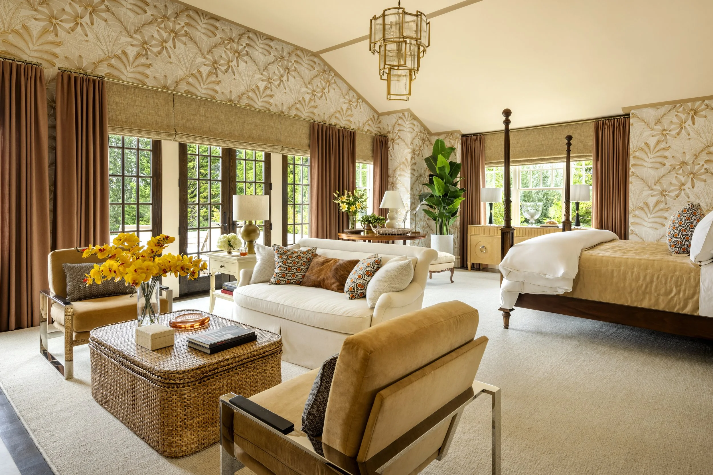 A living room with large windows, beige and brown furniture, floral wallpaper, and various plants and decorative items.