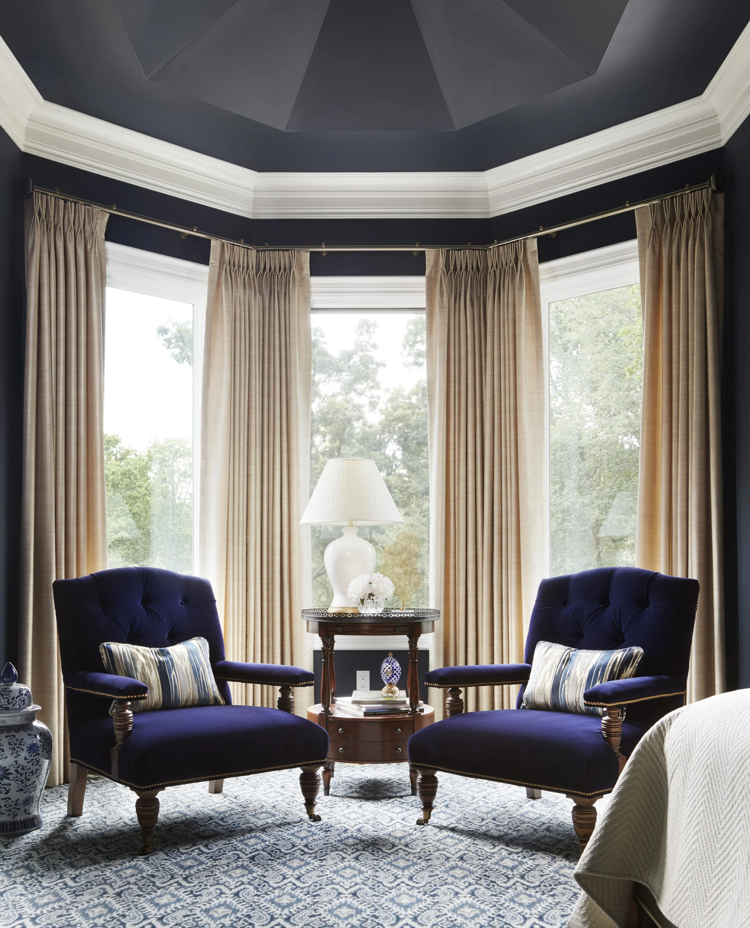 Living room with large bay window covered with beige curtains, two navy blue velvet armchairs with striped cushions, a wooden side table with decorative items, a white table lamp, patterned rug, and landscaped greenery visible outside.