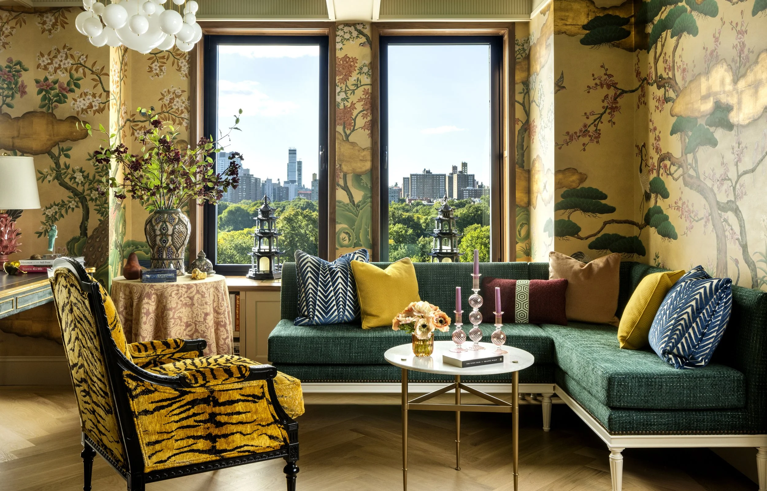 A colorful living room with gold and floral wallpaper, a large window showing a city skyline and green trees, a green sofa with various patterned pillows, a round marble coffee table with pink candles and flowers, a yellow leopard print armchair, wal
